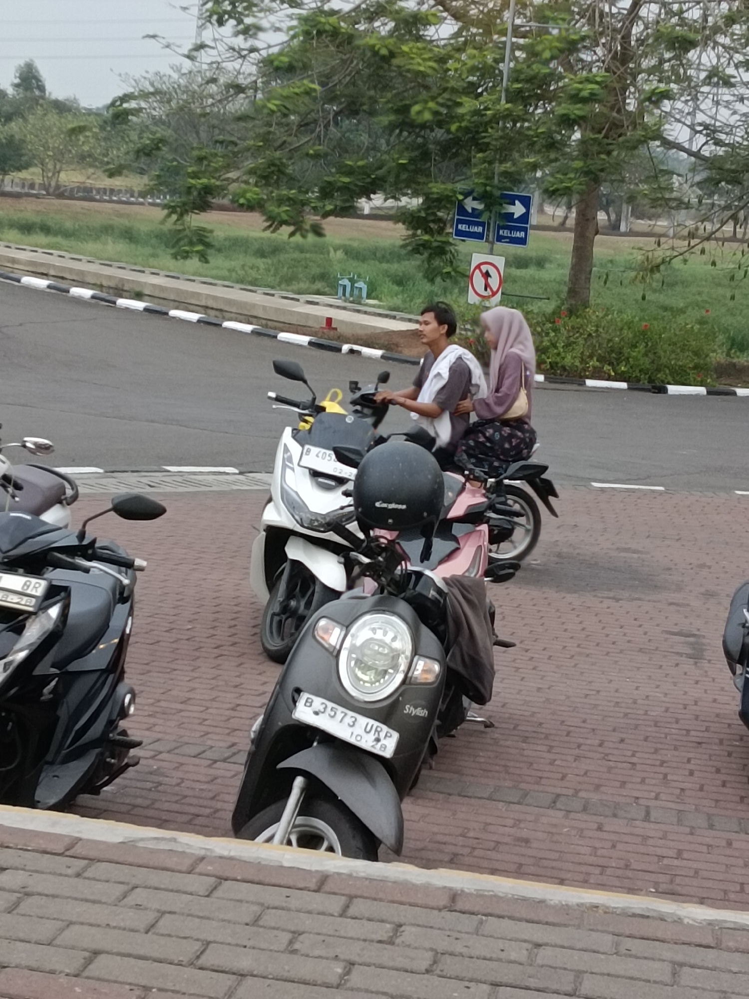 In Jayamukti, Indonesia, a medium shot captures two individuals riding a motorcycle on a paved road. The driver, a male with short dark hair and a light-colored garment draped over his shoulders, is facing forward. The passenger, a female wearing a light purple hijab and a patterned skirt, is seated behind him. Both are positioned on a motorcycle that appears to be moving. Several other motorcycles are parked or in motion in the foreground and background. The environment includes a paved road with white and black markings, a grassy area, trees, and signage indicating directions and prohibitions. The sky is overcast, suggesting a cloudy day. The scene is captured from a slightly elevated perspective, looking down at the motorcycles and riders.