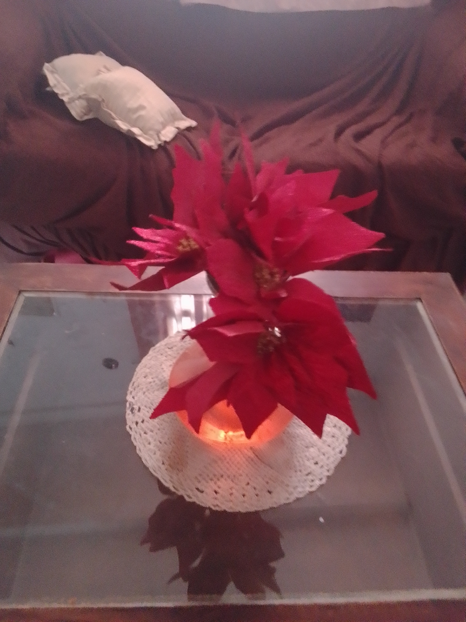 The image captures a dimly lit indoor scene, likely a living room, with a brown sofa in the background and a glass-topped coffee table in the foreground. On the table, a red poinsettia arrangement sits atop a white crocheted doily surrounding a lit orange candle. A light-colored pillow is visible on the sofa. The scene is devoid of any people or discernible activity, suggesting a static, posed shot or a moment captured in time. The overall ambiance is warm and festive, possibly indicative of a holiday setting. No information regarding the location in La Tagua, Venezuela can be derived from the visual content of the image.