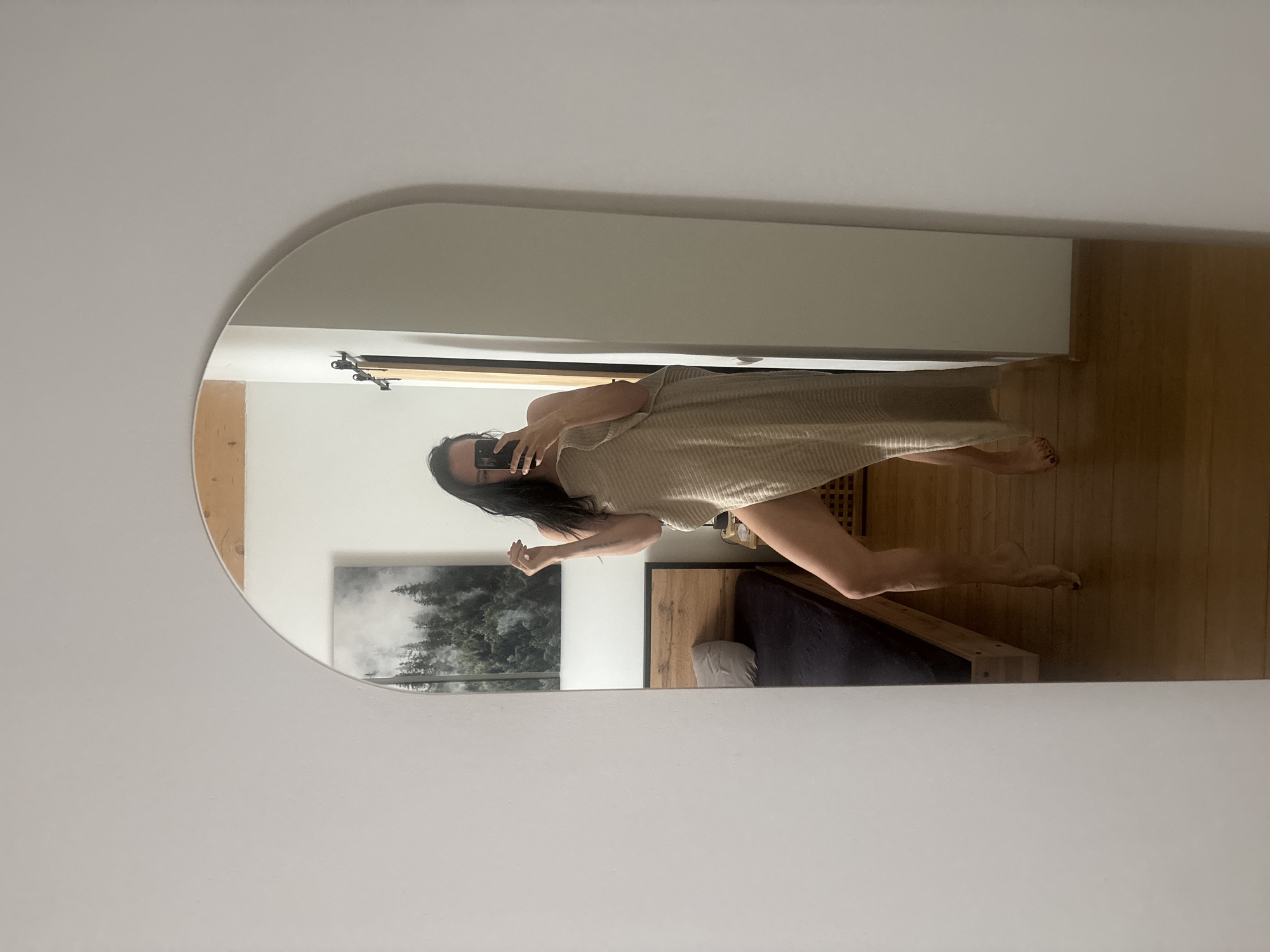 A woman is visible in a mirror, holding a phone and taking a photo of herself. She is wearing a light-colored garment that covers her torso. The room reflected in the mirror contains a bed with a dark blanket and light pillows, wooden floors, and a landscape painting of a forest on the wall. There is no indication of the location in the city of Chaikovsky, Russia.