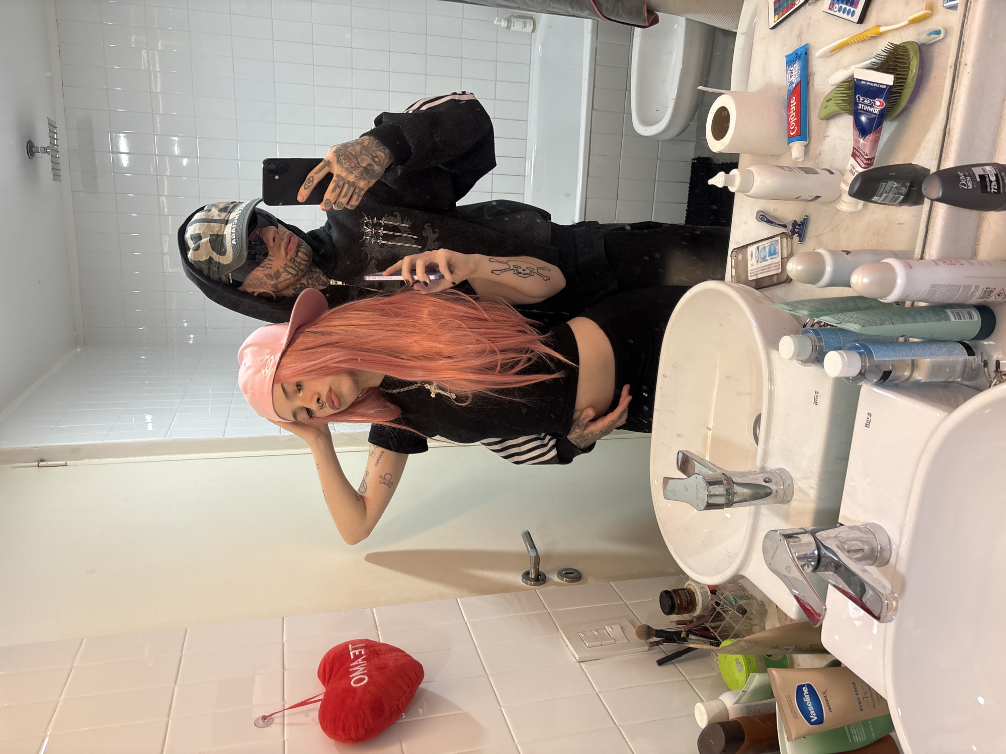A mirror selfie captures two individuals in a residential bathroom in Buenos Aires, Argentina. A female subject, with long pink hair, a pink baseball cap, a black crop top featuring white Adidas stripes, and dark pants, is positioned on the left. She has visible tattoos on her arms and a septum piercing, with her right hand near her cap and her left hand resting on her stomach. Behind her and to the right stands a male subject, clad in a dark hooded sweatshirt with a graphic, a dark baseball cap (showing a reversed Balenciaga logo), and sunglasses. He exhibits extensive facial tattoos, ear gauges, and multiple tattoos on his hands and arms. He holds a black smartphone in his left hand, taking the photograph, while his right arm is around the female's waist. Both individuals are looking towards the camera. The bathroom features white tiled walls, a white door, a dual-sink vanity with chrome faucets, and a countertop cluttered with various toiletries including lotions, deodorants (Dove Men+Care visible), toothpastes (Colgate and Pomada brands visible), a razor, and makeup brushes in a clear holder. A red plush heart with "TE AMO" (Spanish for "I love you") inscribed hangs on the tiled wall via a suction cup. In the background, a white bathtub, a bidet, and a square shower head are visible. A black mat is present on the floor.
