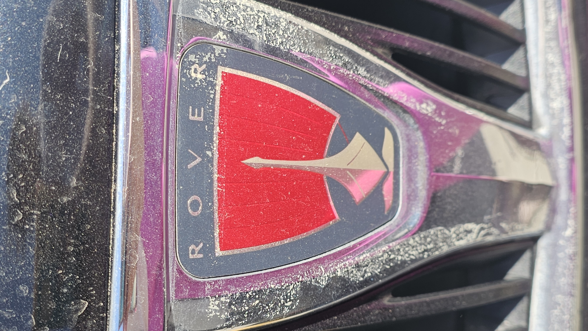 A close-up, vertical photograph depicts a soiled Rover automobile emblem affixed to the front grille of a vehicle. The emblem features the word "ROVER" in silver lettering across its upper dark-colored section, positioned above a red, shield-shaped field that contains a stylized silver depiction of a Viking longship's prow and sail. The emblem's chrome border and the surrounding dark vehicle surfaces are heavily coated with a fine, light-colored layer of dust, dirt, and visible water spots. Minor abrasions and scratches are evident on the metallic trim and the badge's dark background. Pink and magenta reflections are present on the reflective surfaces, indicating ambient light conditions. Vertical slats of the vehicle's dark-colored grille are partially visible below and to the sides of the emblem, also covered in dust. The scene is located in San Bernardino Tlaxcalancingo, Mexico. No individuals, overt actions, or interactions are visible.