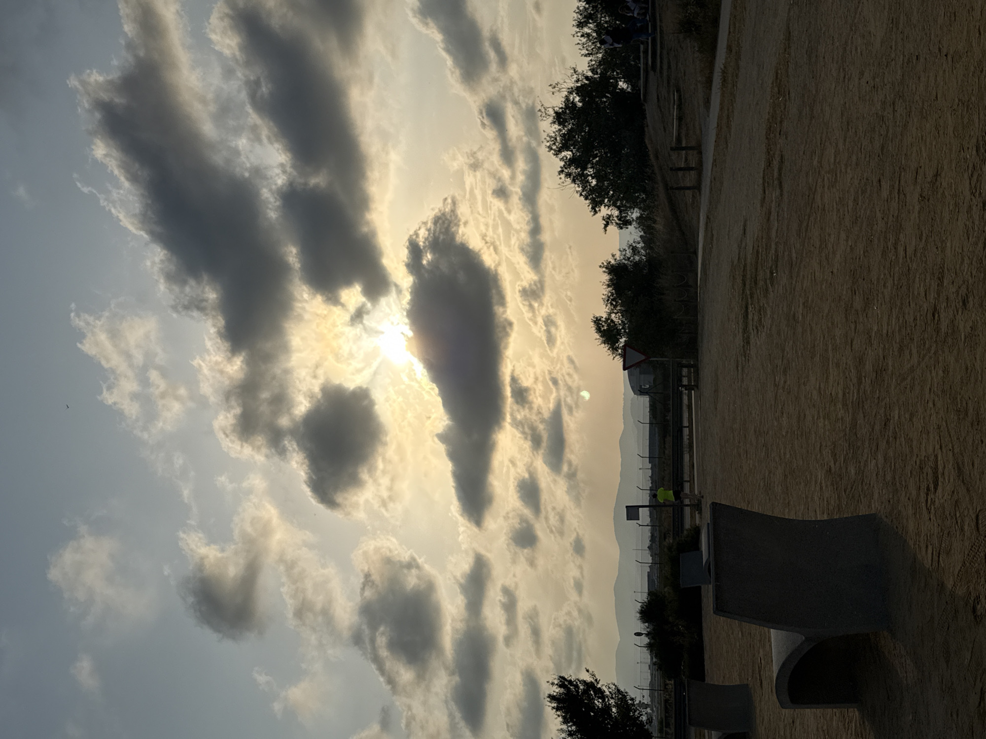 The image depicts an outdoor scene with a focus on the sky and landscape at dusk. The sky is filled with dramatic clouds, some dark and heavy, others illuminated by the setting sun, which is partially obscured by a large cloud formation. The sun's rays create a warm, golden glow across the upper portion of the sky.
In the foreground, a sandy or gravelly ground area is visible, with several modern, curved concrete benches. One bench is prominently featured on the left, casting a shadow. Further back, a path or track leads into the distance. A lone figure in a bright yellow shirt is jogging or running along this path.
The middle ground features a line of trees and vegetation, behind which a fence and some distant structures can be seen. The background consists of a range of hills or mountains silhouetted against the hazy sky. The overall atmosphere is serene and slightly melancholic, characteristic of a sunset in an open area.
The location is identified as a park or open space in El Prat de Llobregat, Spain, likely near the coast or an airport given the distant structures and flat landscape. The presence of the running figure and the benches suggests a public recreational area.