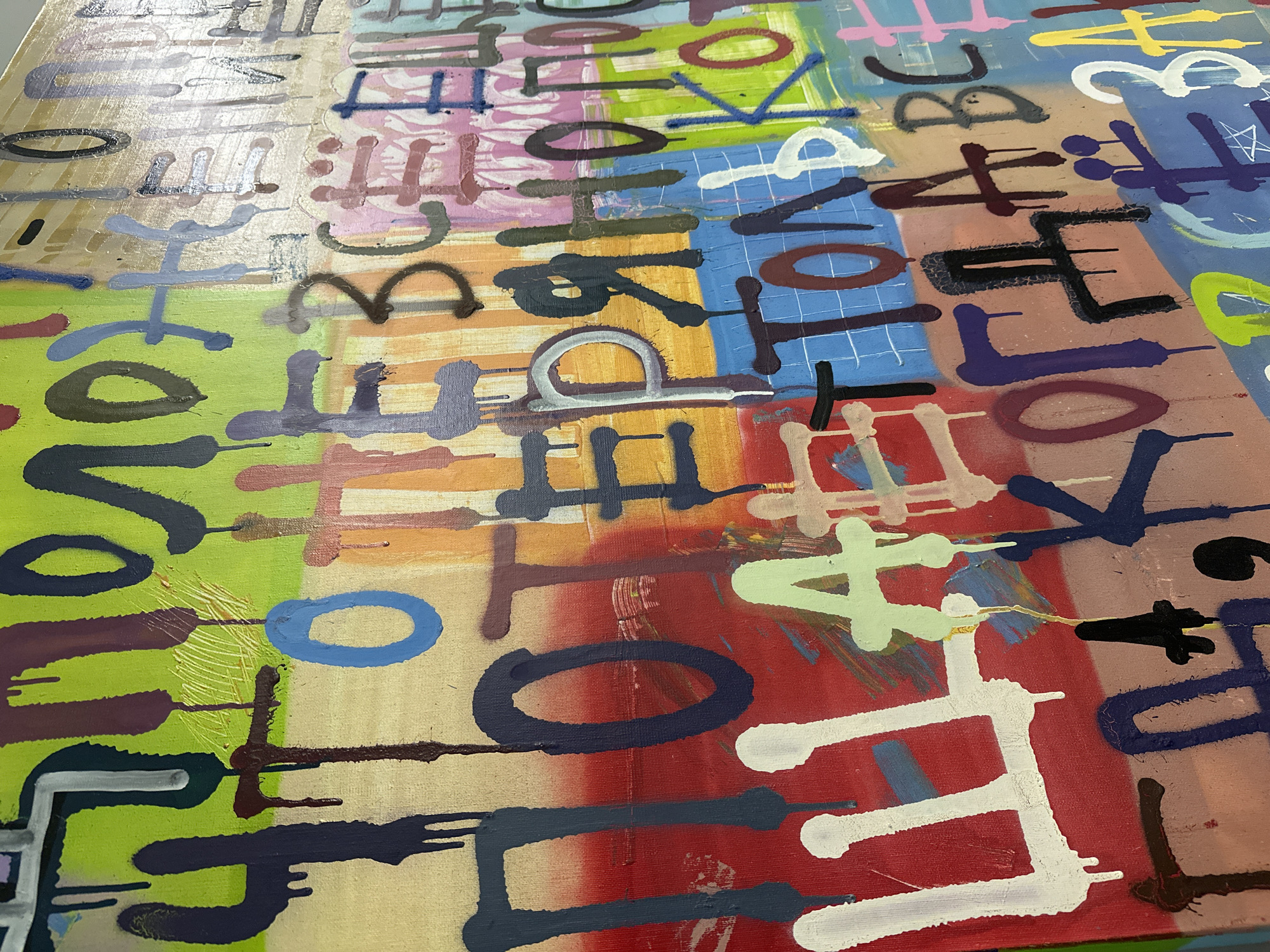 The image depicts an abstract painting with a grid-like background of various colors. Overlaid on this grid are numerous stylized letters and shapes rendered in a variety of colors, including white, black, blue, green, purple, and brown. The style is reminiscent of graffiti or street art. The painting appears to be on canvas, and the image is taken at a close angle, suggesting it may be viewed indoors. There is no indication this artwork is located in Moscow, Russia. No people, subjects, objects, actions, or interactions are indicated from the photograph.