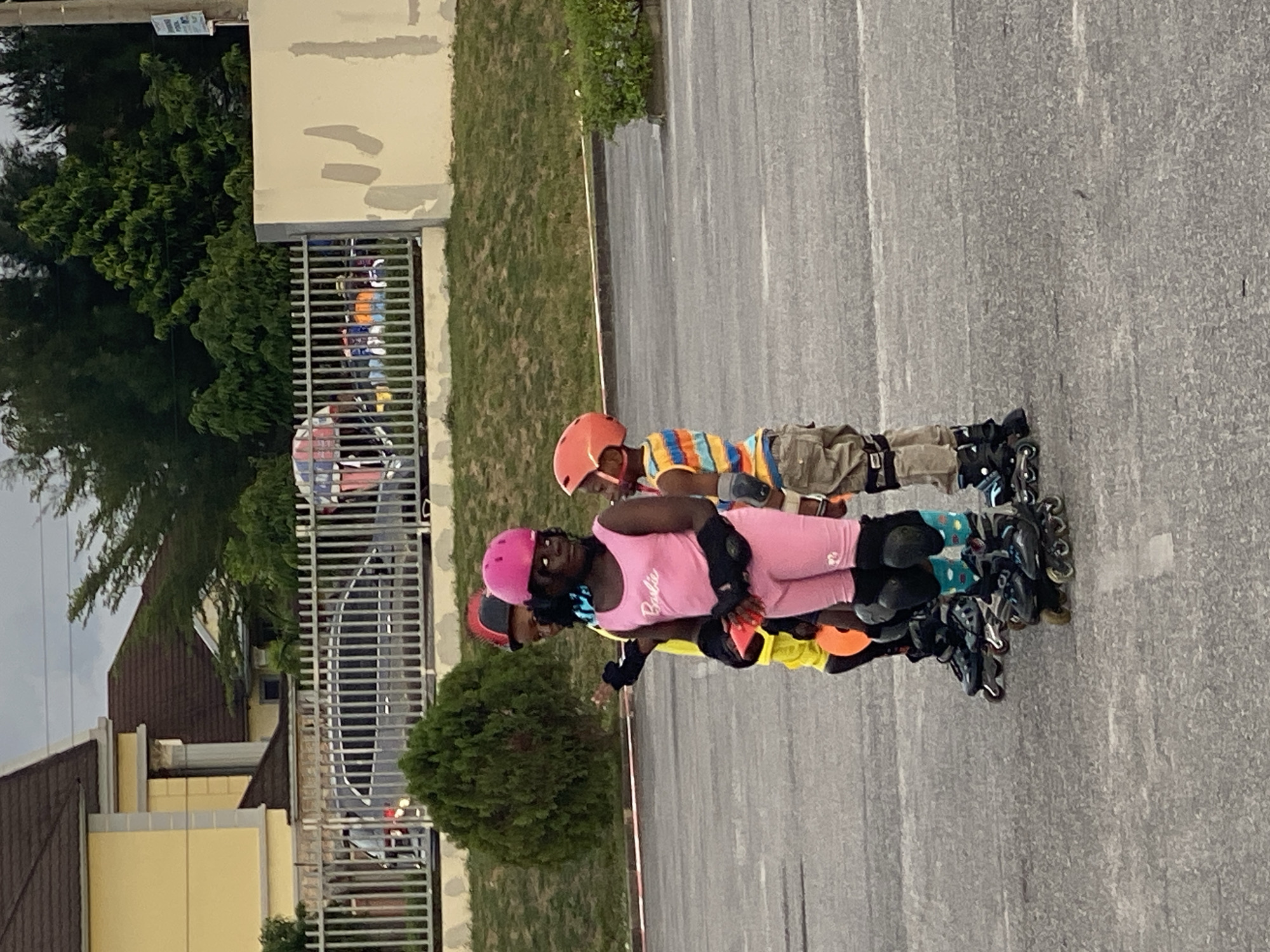 Three individuals are inline skating on a paved surface. The first skater wears a pink helmet and a pink outfit with "Barbie" printed on the shirt, as well as elbow and knee pads. The second skater wears a yellow top and an orange helmet. The third skater wears a striped shirt, brown pants, and an orange helmet. They all wear inline skates. The background includes a building, a fence, and some greenery. The image was taken in Sangotedo, Nigeria.
