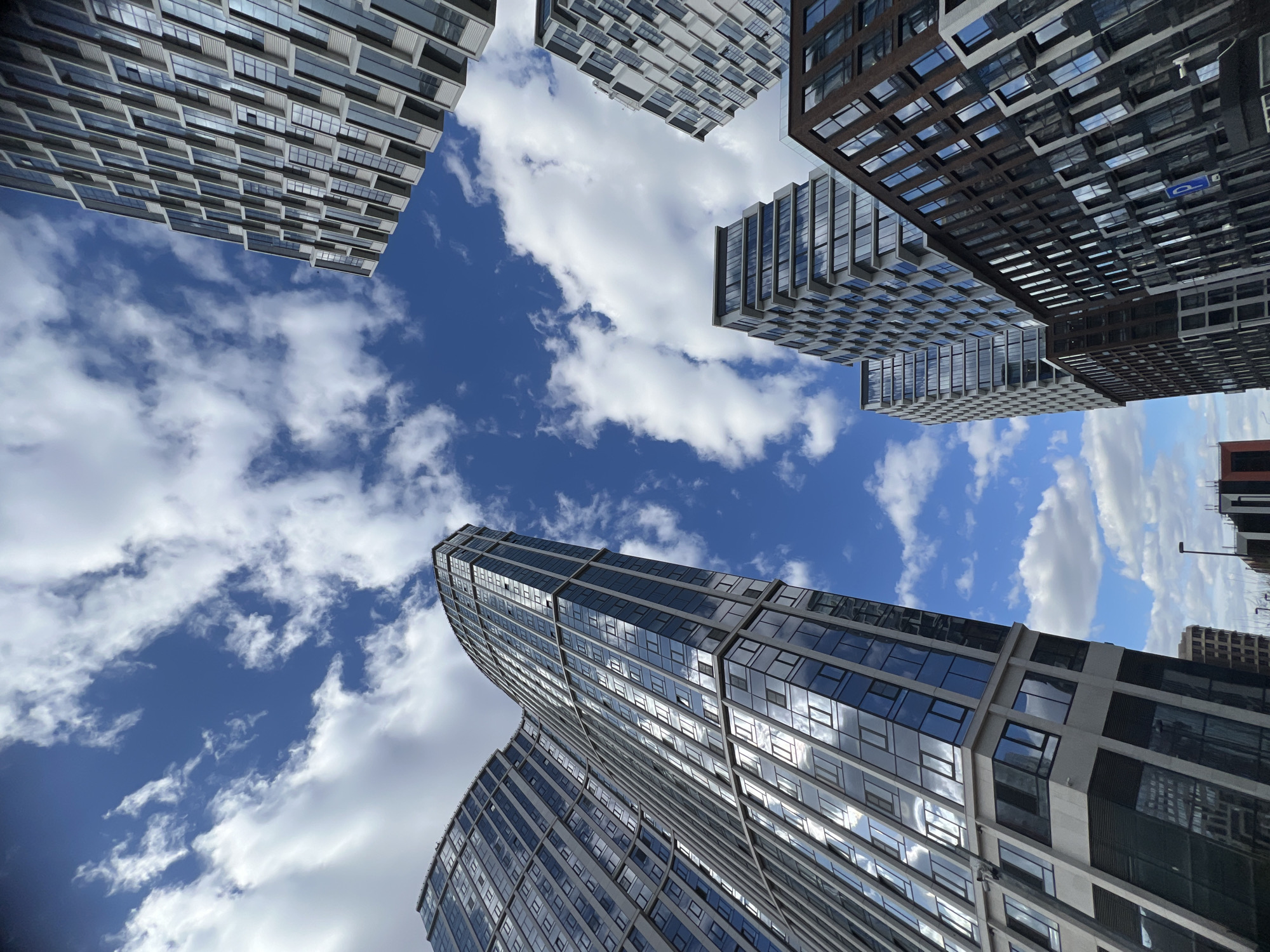 Looking upwards, the image captures several modern buildings against a backdrop of a blue sky dotted with white clouds. The architecture is contemporary, featuring glass windows that reflect the sky and clouds, and the building materials range from light to dark shades. The image suggests a location within a city with modern high-rise buildings, potentially Khimki, Russia. The structures surround the viewer, creating a sense of urban density.