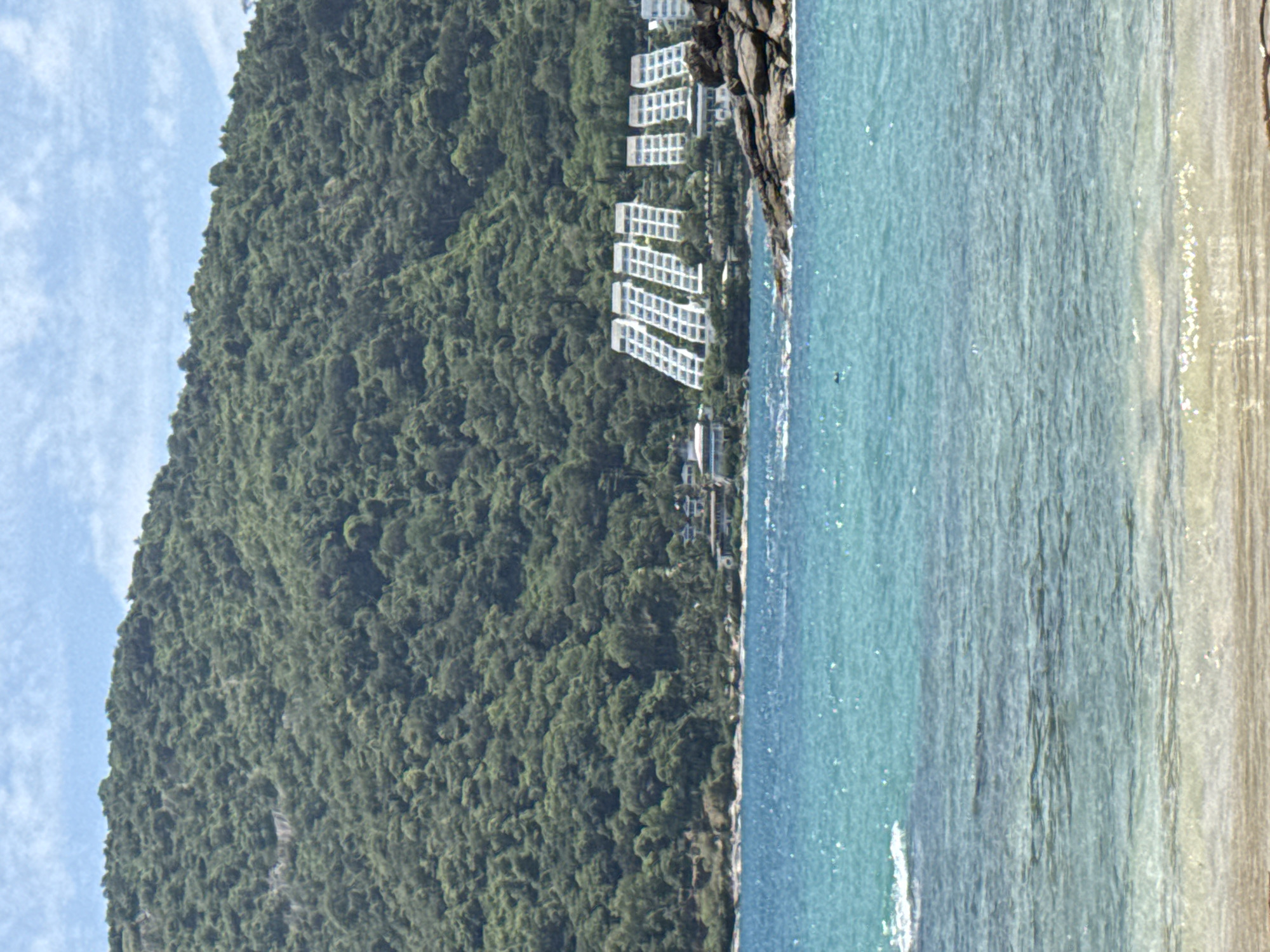 The image captures a coastal scene in Rawai, Thailand, featuring a lush, tree-covered mountain that dominates the background. Nestled at the foot of the mountain, along the shoreline, are several modern, tiered white buildings with a repetitive architectural design. The buildings appear to be resorts or hotels, arranged in rows and facing the sea. The turquoise water in the foreground is clear, with some waves gently lapping at a sandy beach. Rocks are visible along the shoreline, and the sky is a light blue with scattered white clouds. The overall impression is one of a serene tropical location.