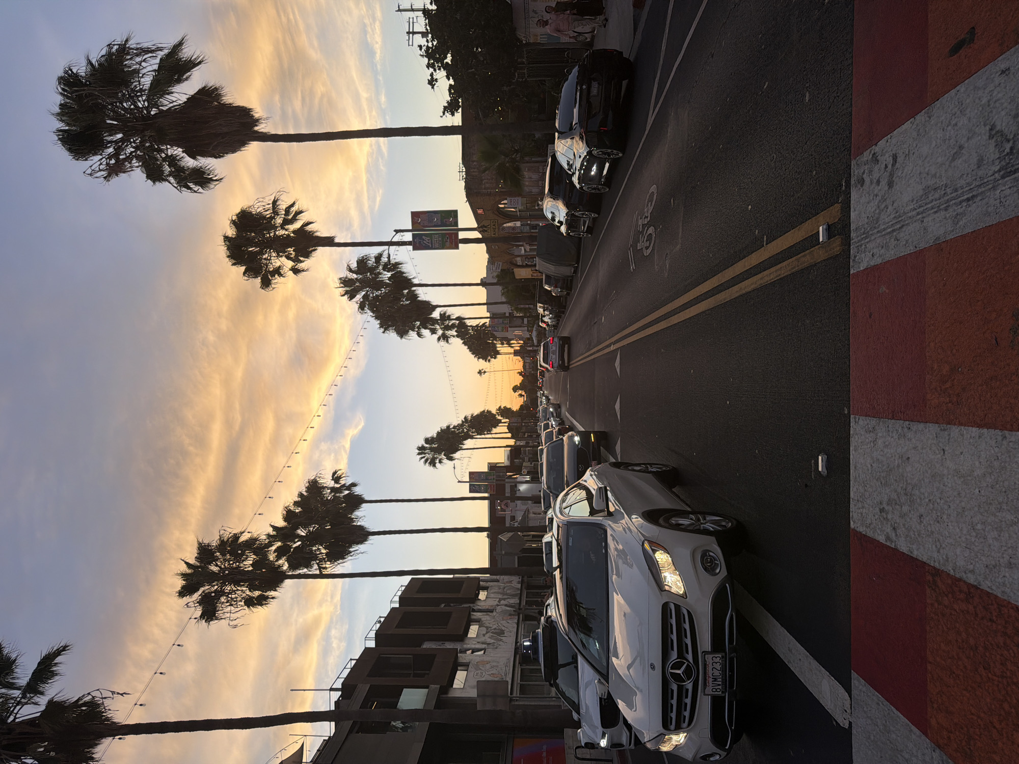 A street scene in Los Angeles, United States of America, at sunset, features a traffic jam on a wide, paved road lined with palm trees. The sky displays an array of warm colors from yellow to orange and purple, with thin clouds stretching horizontally. Streetlights are strung between poles along the road.
In the foreground, a white Mercedes-Benz GLA appears to be stationary in traffic, with its headlights on. To its left, a white Jaguar is also stopped. A red and white pedestrian crossing marks the immediate foreground.
Further down the street, the traffic continues with various cars, including a black sedan with distinctive rims. Buildings of varying architectural styles line both sides of the road. On the left, a modern building with dark facade and balconies stands next to a lower structure with a mural on its wall. On the right, a building with arched doorways and a sign reading "ART AHEAD" is visible.
Several individuals are seen walking on the sidewalk on the right side of the street. The overall atmosphere suggests a typical evening in a bustling urban environment during a scenic sunset.