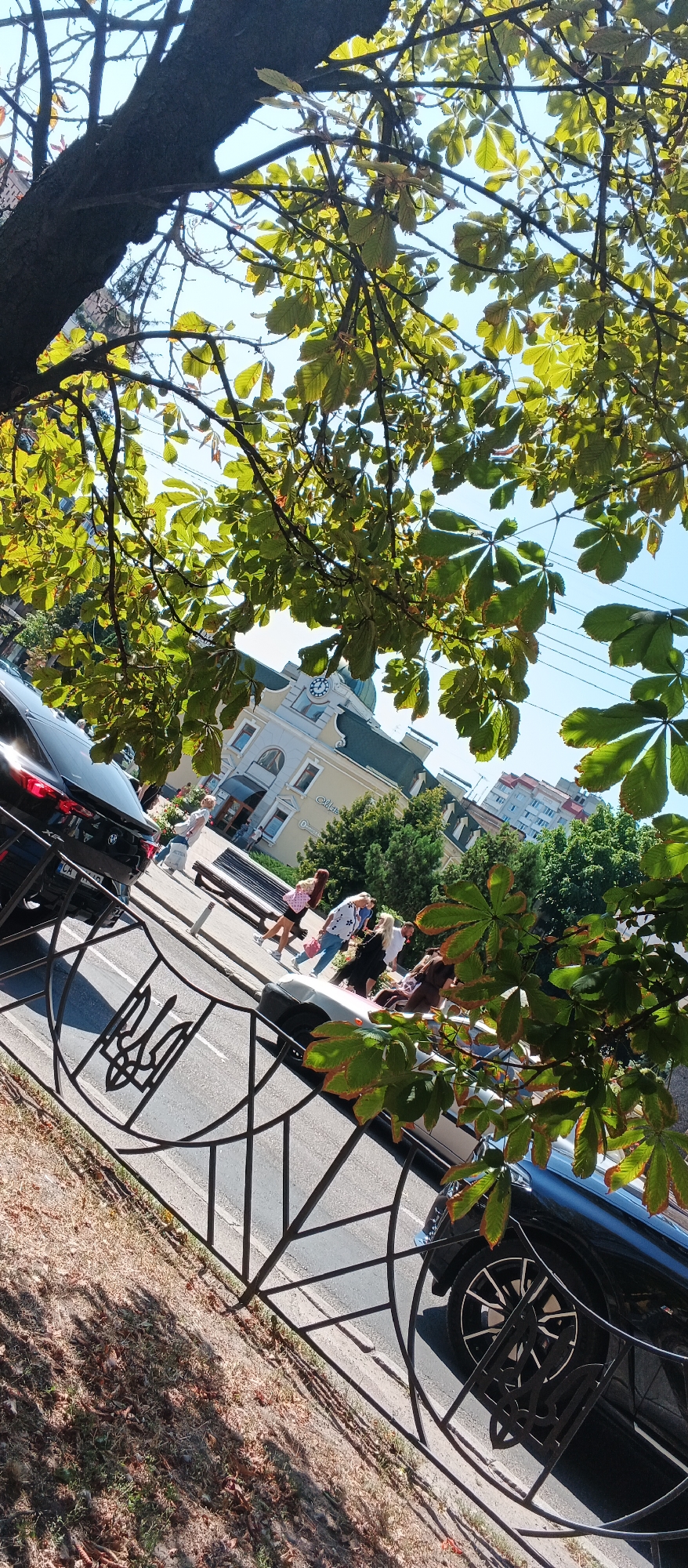 The image shows a street scene in the city of Cherkasy, Ukraine, on a sunny day. A black SUV is parked on the left side of the road, partially obscured by tree branches. In the background, a building with a clock tower is visible, and several people are walking on the sidewalk. A metal fence with a trident emblem runs across the foreground. The overall scene is that of a typical urban environment with elements of nature and public space.