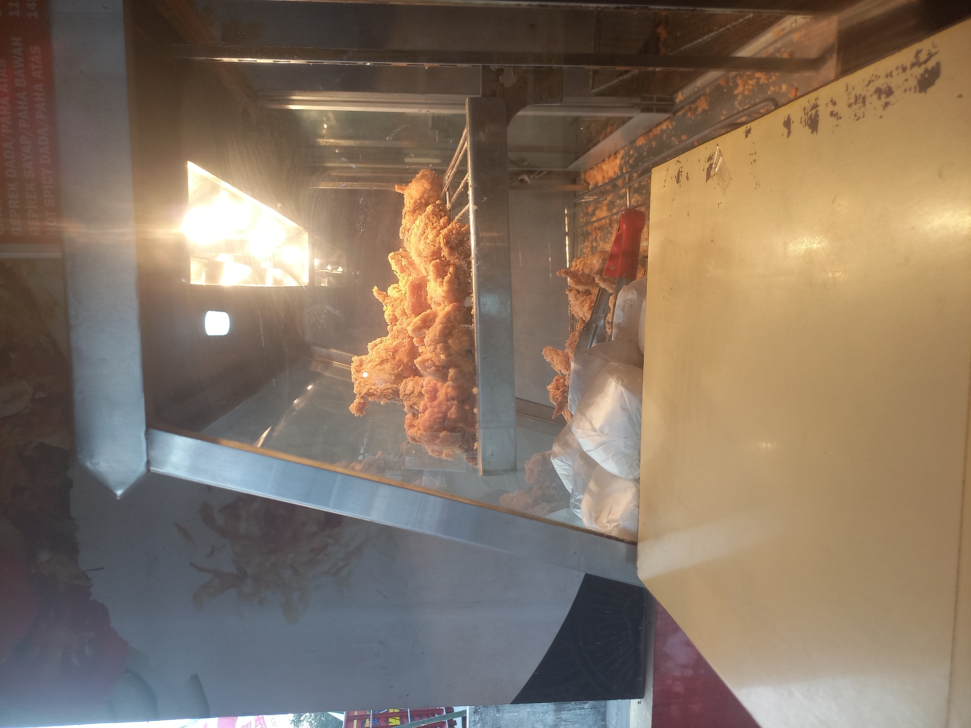 The image shows a close-up view of a display case filled with fried chicken. The fried chicken is golden brown and appears to be crispy. Some of the chicken pieces are whole, while others are cut into smaller pieces. The display case is made of glass and metal, and it is lit by bright overhead lights. In the foreground, there is a beige-colored counter or table with some smudges and debris on its surface. There are also some white paper bags and a red plastic utensil visible in front of the display case. The background is blurred, but it appears to be an indoor setting, possibly a restaurant or food stall. The text visible on the wall behind the display case suggests it is a food establishment in Bedah Lawak, Indonesia, with a menu listing various chicken dishes.