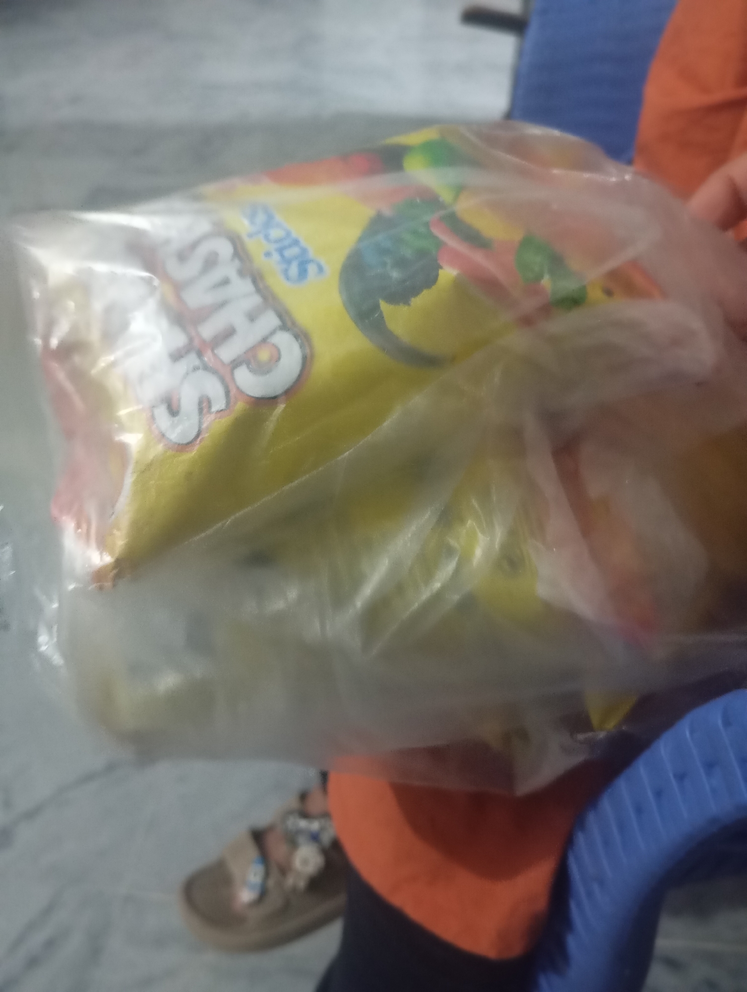 A close-up, blurry image shows a person holding a clear plastic bag containing a yellow-labeled snack with the word "CHAS" partially visible in large, bold white letters. The word "Sticks" is also visible in blue lettering. The snack package has colorful graphics, including what appears to be a cartoon dinosaur or lizard. The background is out of focus, but suggests an indoor environment with a tiled floor and hints of furniture, possibly in Lassan, Pakistan. The person's hand, wearing a sandal with floral embellishments, is visible at the bottom left.
