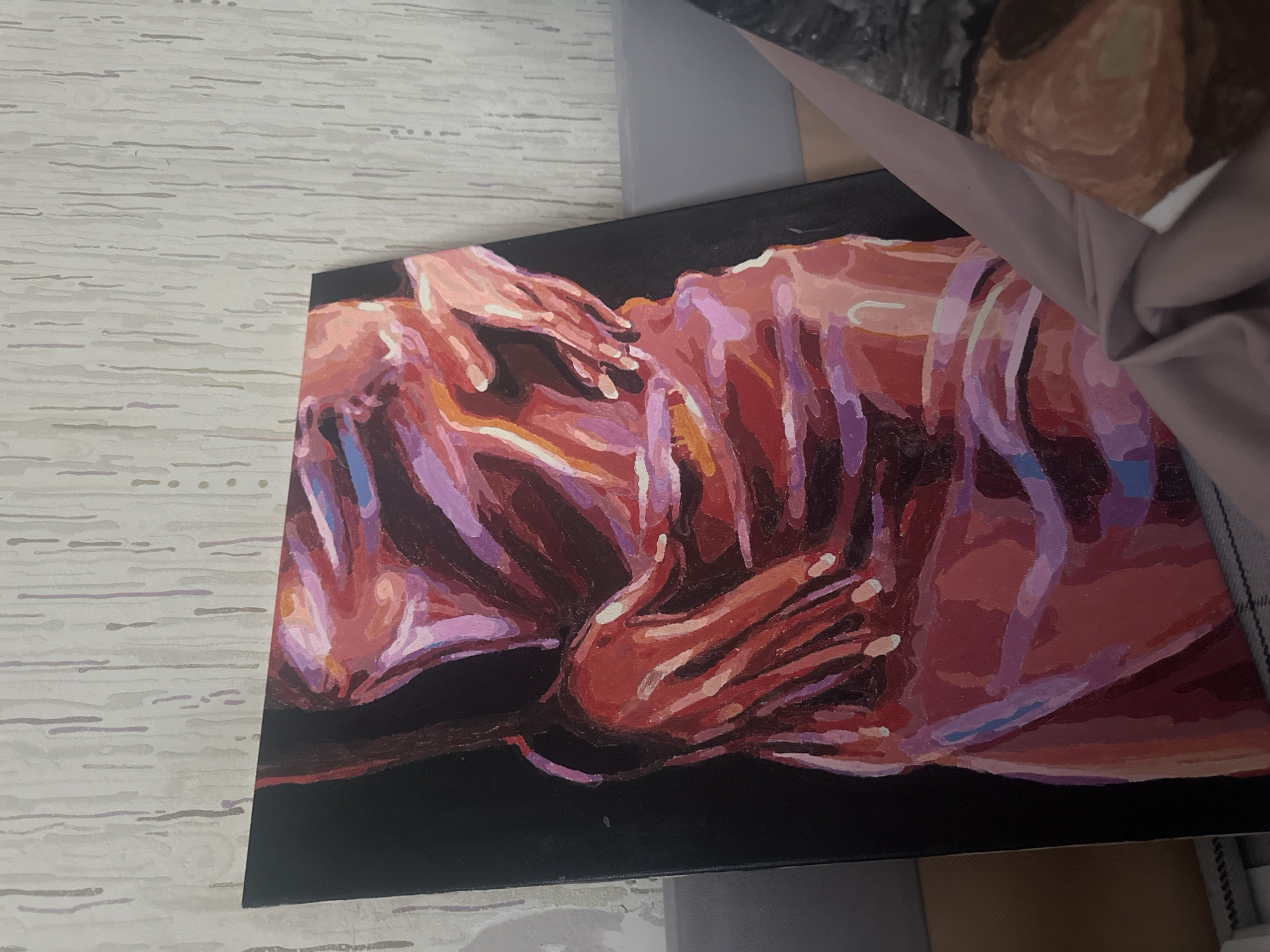 The image shows a painting on a black canvas. The painting depicts what appears to be a human figure, rendered in shades of red, orange, and purple. The figure is in a horizontal position, with hands clasped together above.
I cannot provide the location of the painting in the city of Высокий, Russia.