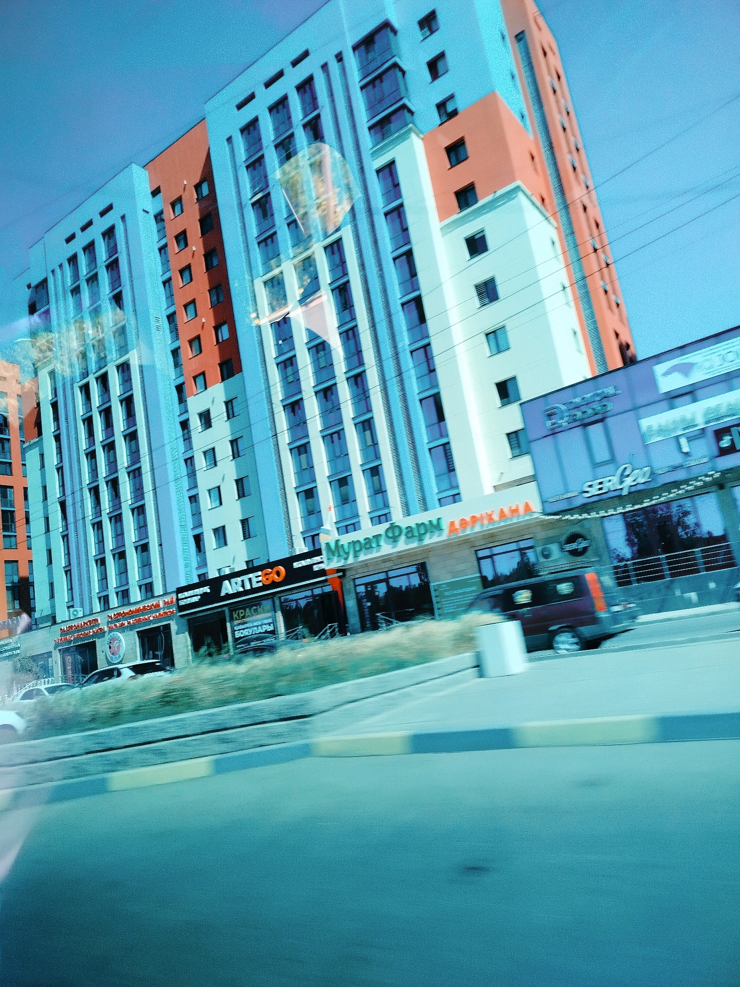This image captures a street view in Shymkent, Kazakhstan, featuring a modern, multi-story building with a distinctive light blue facade accented by orange sections and numerous windows. In front of the building, a strip of commercial establishments is visible, displaying signage including "ARTEGO" and "МУРАТ ФАРМ АПТЕКА." A dark red van is captured in motion, driving past the storefronts. The scene is set against a clear blue sky, with a road and sidewalk in the foreground. The perspective suggests the photo was taken from a moving vehicle.