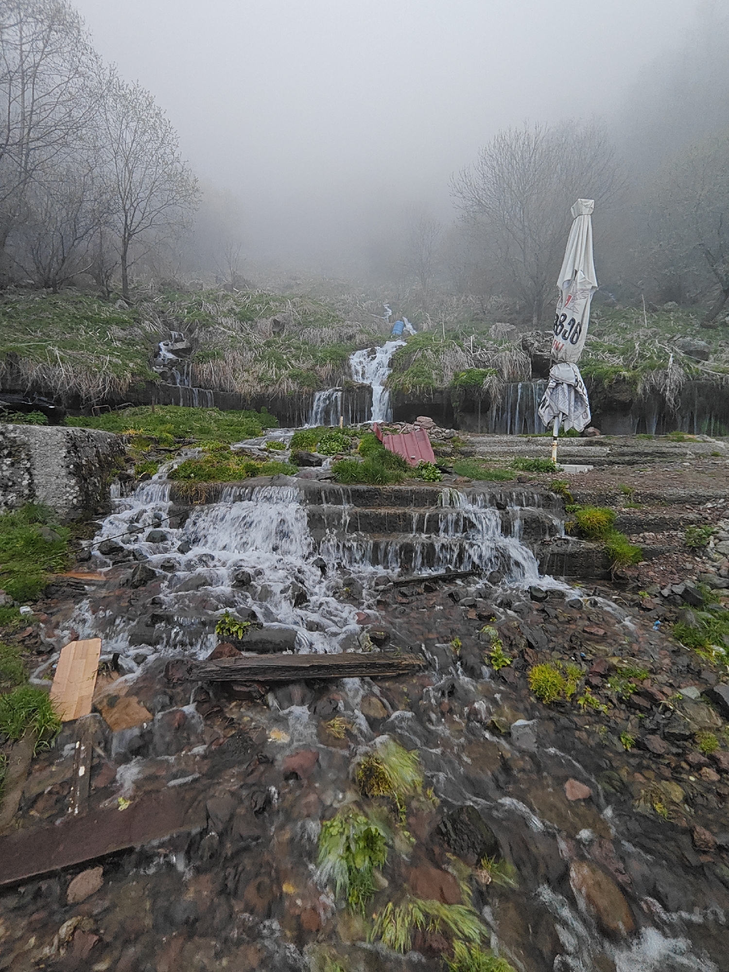 This media file depicts a misty, damp natural landscape in Zemo Mleta, Georgia, dominated by a cascading stream. The scene is overcast, with dense fog or mist obscuring the background, where bare trees with emerging leaves are visible as muted silhouettes.
In the foreground, a lively stream flows down a series of small waterfalls and rapids, navigating over rocks and what appear to be constructed steps or ledges. The water is clear and white-foamed as it tumbles. Various debris, including wooden planks, rests in and alongside the stream. Patches of vibrant green grass and moss cling to the rocks and soil along the water's edge.
Mid-ground, to the right of the stream, a large, folded white umbrella stands upright. Black text is visible on the umbrella, clearly showing "3336" and some partially obscured lettering above it. Near the umbrella, a reddish, corrugated sheet lies on the ground. Further up the slope, a blue barrel is nestled amongst the vegetation next to the stream's higher reaches. The terrain consists of stepped banks, covered in grass and rocks, leading up into the mist.
No people are visible in the image. The atmosphere is quiet and cool, indicative of an overcast day, possibly in spring or autumn, given the state of the trees and the heavy mist.