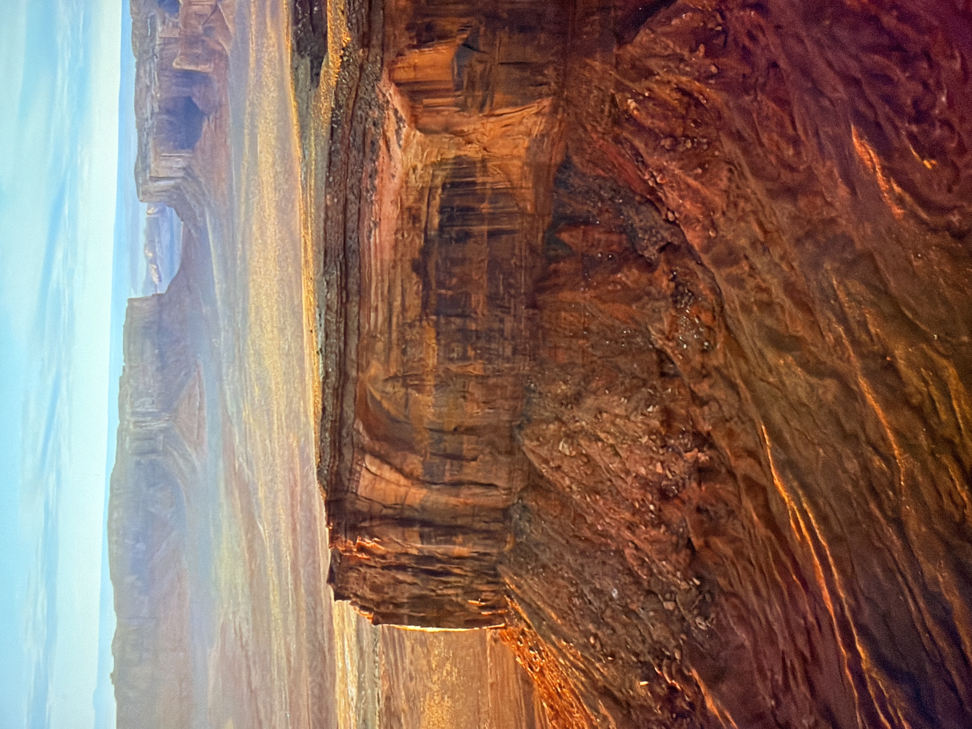 This is a high-angle, full shot of a vast, arid landscape under a bright, clear sky. The scene is dominated by massive rock formations, characterized by layered, stratified cliffs and mesas. The foreground features a steep, rocky incline leading to the edge of a prominent cliff face. This cliff is a deep orange and red, with distinct horizontal striations and vertical streaks, suggesting erosion and water damage. The sunlight casts long shadows and highlights the textured surfaces of the rocks, creating a dramatic interplay of light and dark.
In the middle ground, a wide, flat expanse stretches out, dotted with smaller rock formations and mesas that recede into the distance. These formations are also layered and appear to be composed of similar reddish-brown rock. A hazy atmosphere softens the details of the distant landscape, creating a sense of depth and scale.
The background consists of rolling hills and distant, larger mesas, all rendered in muted tones of brown and gray, fading into the horizon. The sky above is a pale blue, with thin, horizontal clouds. The overall impression is one of rugged, geological grandeur in a desolate, sun-drenched environment. There are no people, animals, or man-made structures visible in the image. The location is not in Kyiv, Ukraine.