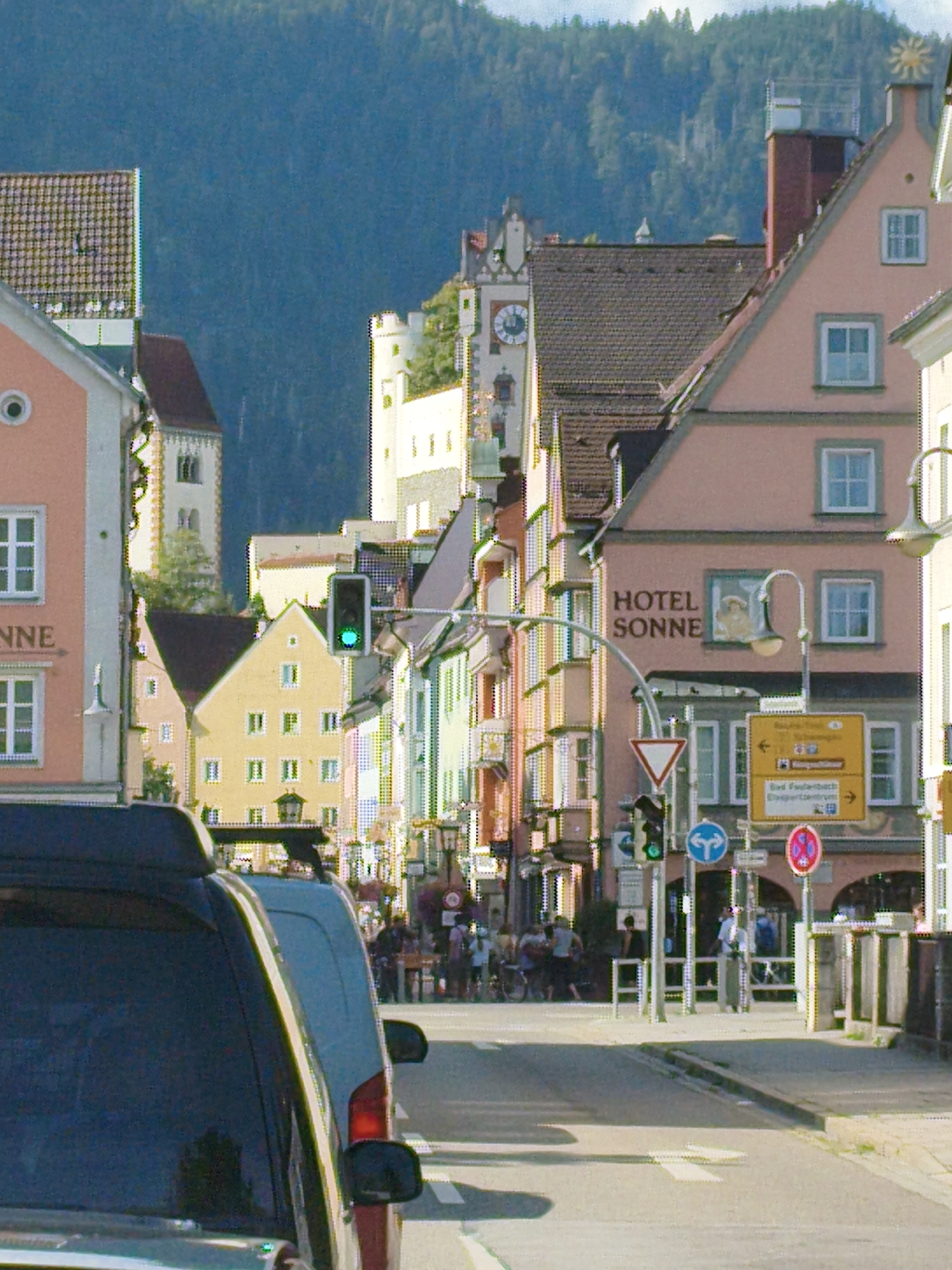 The image depicts a street scene in Füssen, Germany, with a focus on a building labeled "HOTEL SONNE." The building is painted a light pink and has several windows. To the left of the hotel, another building with a tiled roof and circular window is visible. Across the street, a yellow building with multiple windows stands. A traffic light is showing green, and various road signs are present, including directional signs and a circular red and white sign. People are seen walking and standing on the sidewalk, and two vehicles are parked on the street in the foreground. In the background, a densely forested mountain slope is visible under a bright sky.