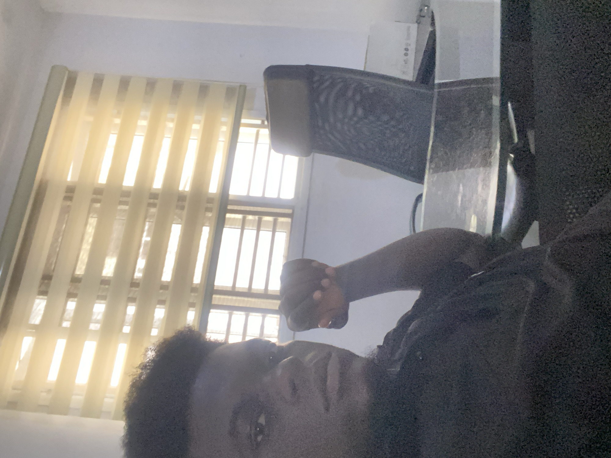 An indoor scene captured during daytime features a person with dark skin and dark, curly hair in the foreground, looking directly at the camera. The image is strongly backlit by a window, causing the person's face and much of their body to appear somewhat shadowed and underexposed. The person's right arm is raised, with their hand clenched into a fist above their head.
Behind the person, a dark office chair with a mesh back is visible. To the left, a large window is covered with translucent vertical blinds, through which bright sunlight streams, illuminating the room and creating vertical stripes of light. The window itself has a visible grid structure. To the right of the person, a reflective surface, likely a desk or table, occupies part of the foreground. In the upper background, behind the chair, a white rectangular object, possibly a box, rests on an unseen surface. The overall lighting creates a high-contrast atmosphere. There is no visible text.