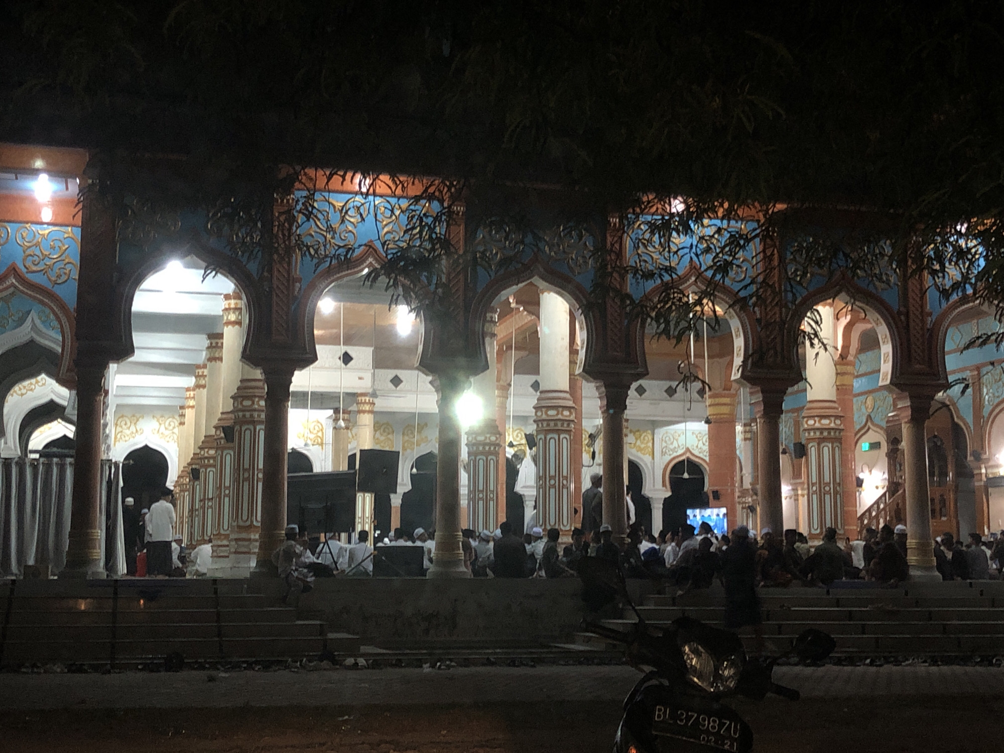 This media file depicts a scene at night, likely after sunset, in front of a mosque in Palohgadeng, Indonesia. The mosque's architecture is characterized by its ornate arches, colorful tiles in blue and gold, and intricately decorated pillars. The time of day is evident from the artificial lighting within the mosque and the overall dark ambiance, with some ambient light filtering through the trees in the foreground.
The primary subjects are a group of people gathered outside the mosque. They appear to be a mix of men and possibly some children, dressed in traditional attire, including caps and sarongs. Some are seated on steps, while others are standing or walking. The presence of a large screen, possibly for displaying religious content or announcements, is visible on the left. Two large speakers are also positioned on the left side of the steps. The gathering suggests a religious event or a community assembly taking place at the mosque.
The foreground is dominated by a motorcycle, which is partially visible, with a license plate reading "BL3798ZU 02.21". The presence of the motorcycle, along with the people gathered, suggests a common, local setting. The overall mood of the scene appears peaceful and communal, with people engaging in religious or social activities under the warm glow of the mosque's lights. The weather appears calm, as there are no visible signs of rain or strong winds. The dense foliage in the upper portion of the image adds a natural element to the urban setting, suggesting a location with both man-made structures and natural greenery.