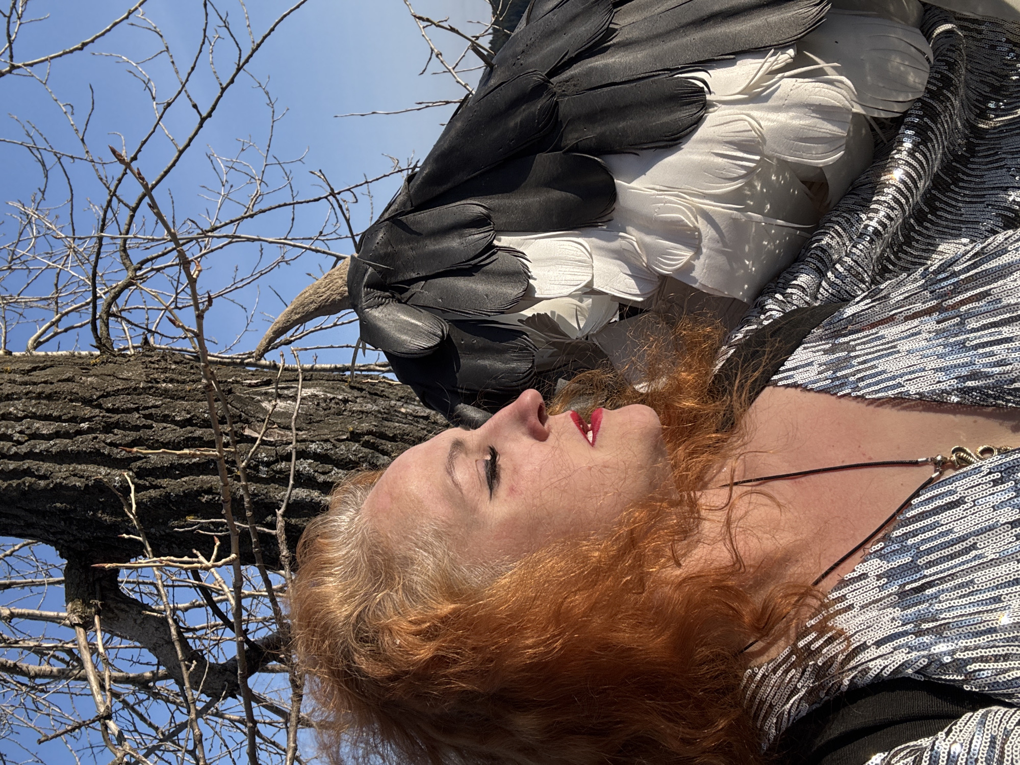 A woman with red hair, red lipstick, and a sequined top lies on her side near a tree branch under a blue sky. Positioned above her is a decorative bird with black and white feathers. The location is not identifiable as Noviy, Russia.