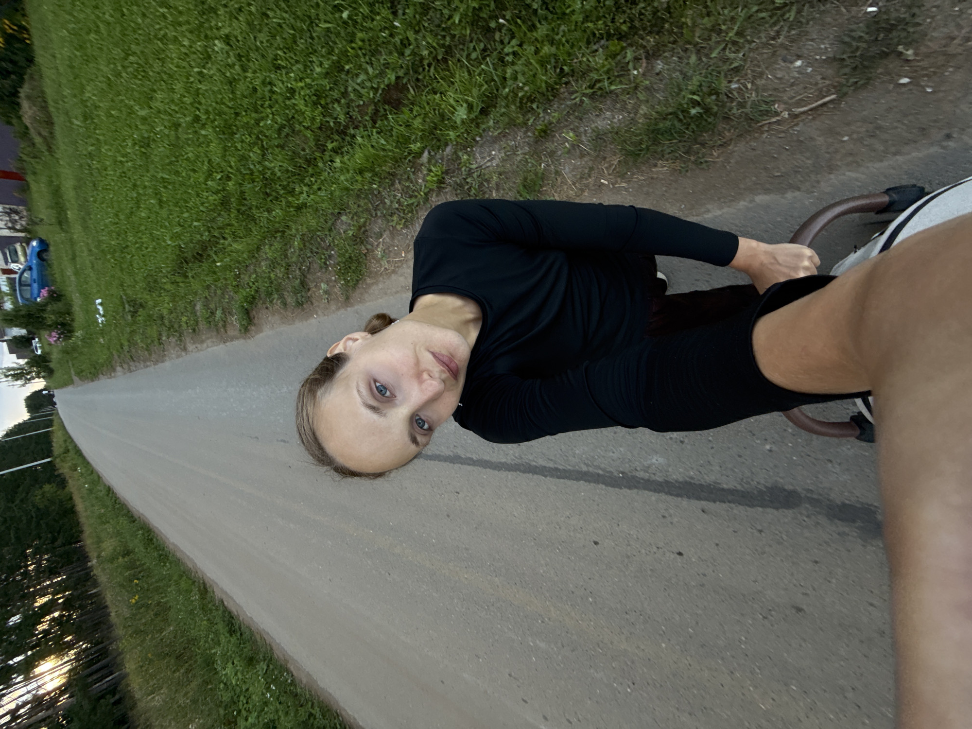 A woman with blonde hair pulled back is taking a selfie while holding the handle of a stroller on a dirt road. She is wearing a black long-sleeved shirt and appears to be outdoors in a rural area. The road is flanked by green grass and trees on the left, and grass and some houses on the right. A blue car is parked in the background on the right side of the road. The sun appears to be setting, casting long shadows from the trees. The location is identified as Podshivalovo, Russia.
