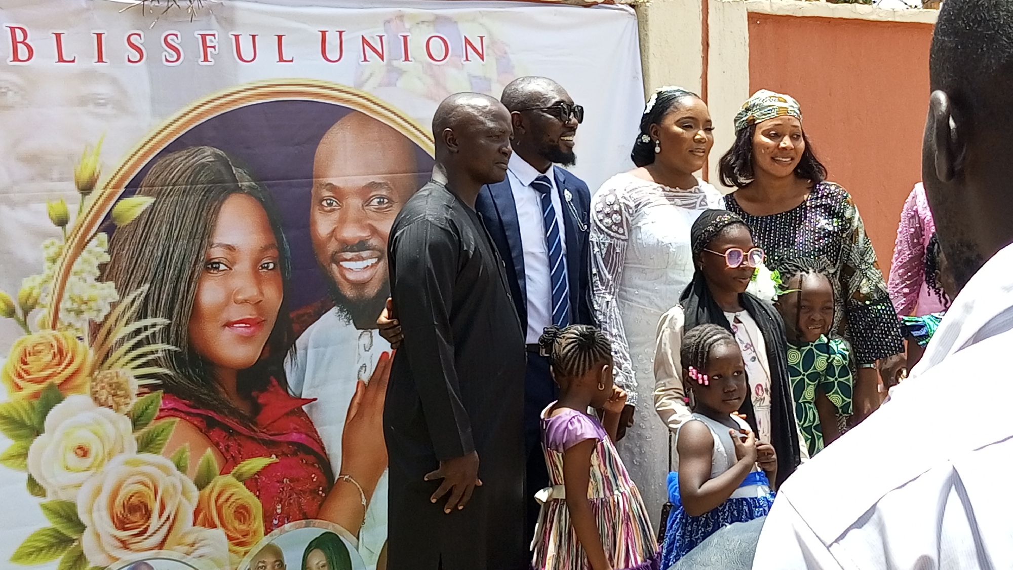 A daytime outdoor scene in Kakau, Nigeria shows a group posing in front of a "Blissful Union" banner. The banner features a couple's photo surrounded by roses. To the right of the banner, three men stand, two in suits and one in traditional attire, flanking a woman in a white dress and another woman in a patterned headwrap and dress. In front of the adults are four young girls in dresses. An individual partially blocks the view from the right. A building is visible in the background.