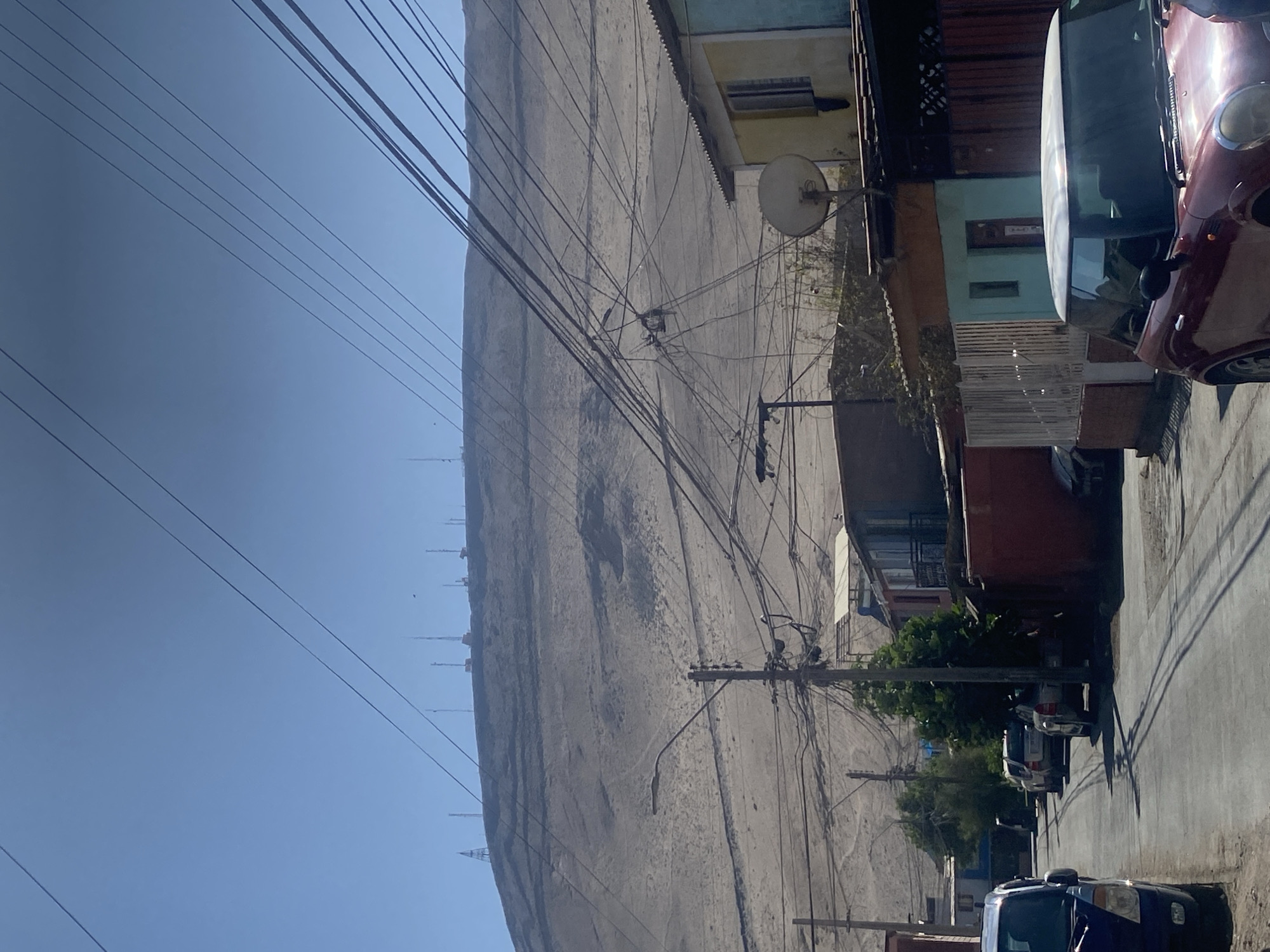 The image shows an urban scene in Arica, Chile. A steep, arid hill dominates the background, heavily scarred with visible lines that may be erosion or previous construction. The foreground features a narrow street lined with densely packed buildings of varying colors and materials. Utility poles and wires are prominent, creating a complex network overhead. Parked cars are visible along the street, with a blue van in the immediate foreground on the right. A satellite dish is attached to one of the buildings. The sky is clear and blue.