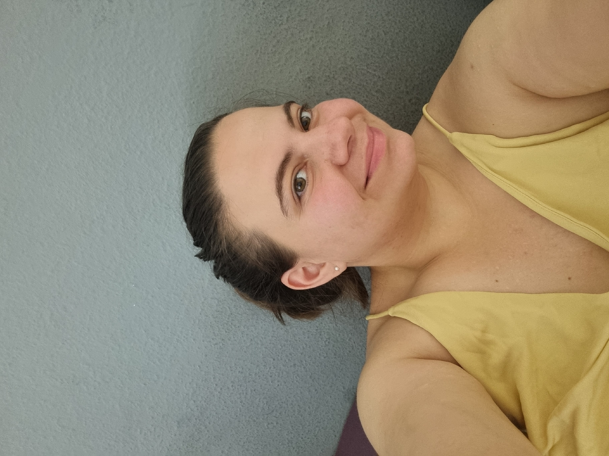 A selfie photograph shows a woman with fair skin and brown hair pulled back away from her face. She is facing toward the viewer with a slight smile. She is wearing a yellow tank top with thin straps. A wall of textured, light-blue material is behind her. The image doesn't provide enough information to place the subject in Saarbrücken, Germany.