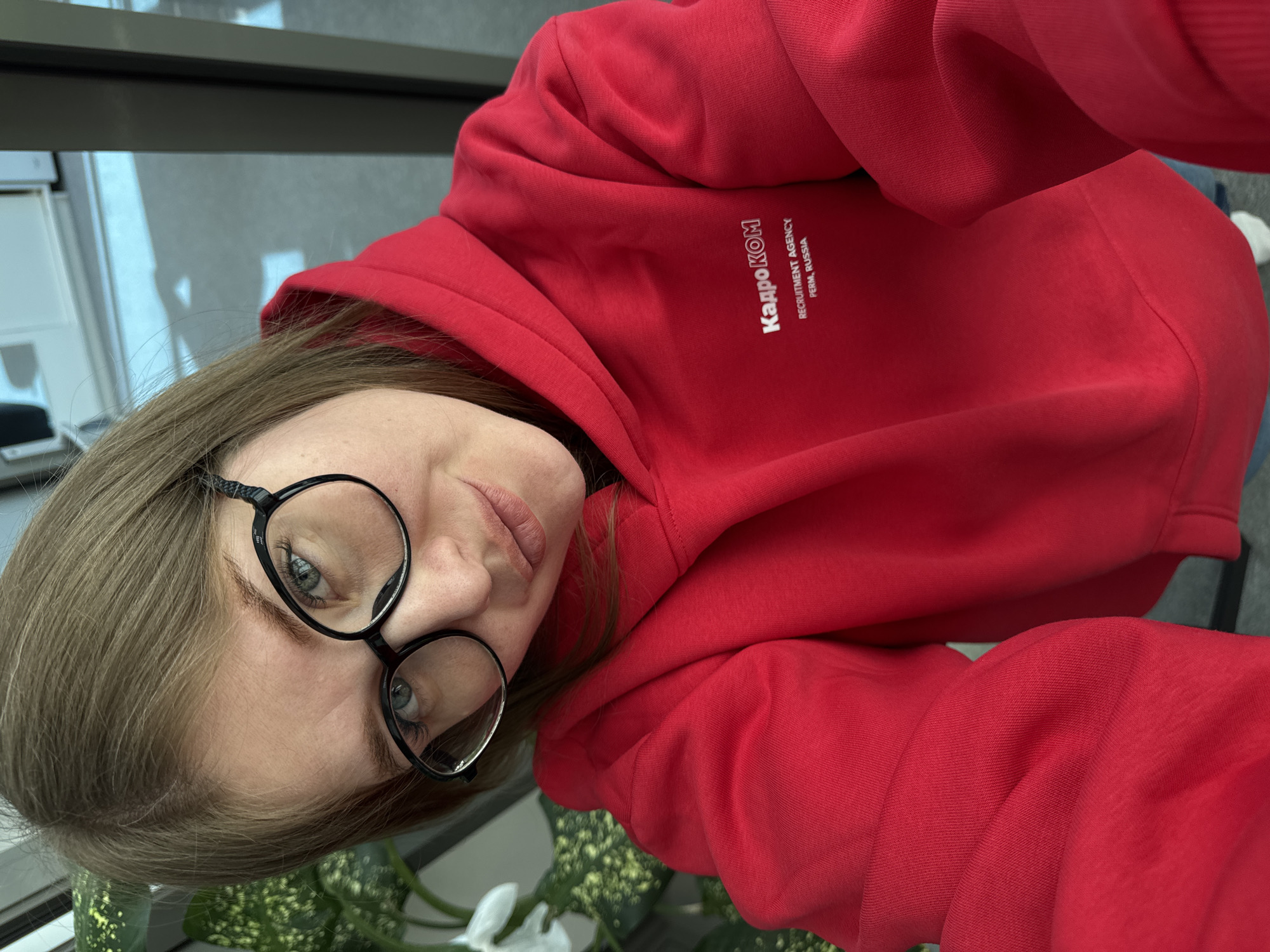 A fair-skinned person with shoulder-length brown hair and round black glasses is taking a selfie. They are wearing a red hooded sweatshirt with white text in Russian that reads "КАДРОКОМ RECRUITMENT AGENCY PERM, RUSSIA". The background appears to be an office setting, with windows and plants visible. The location is Perm, Russia, as indicated by the text on the sweatshirt.