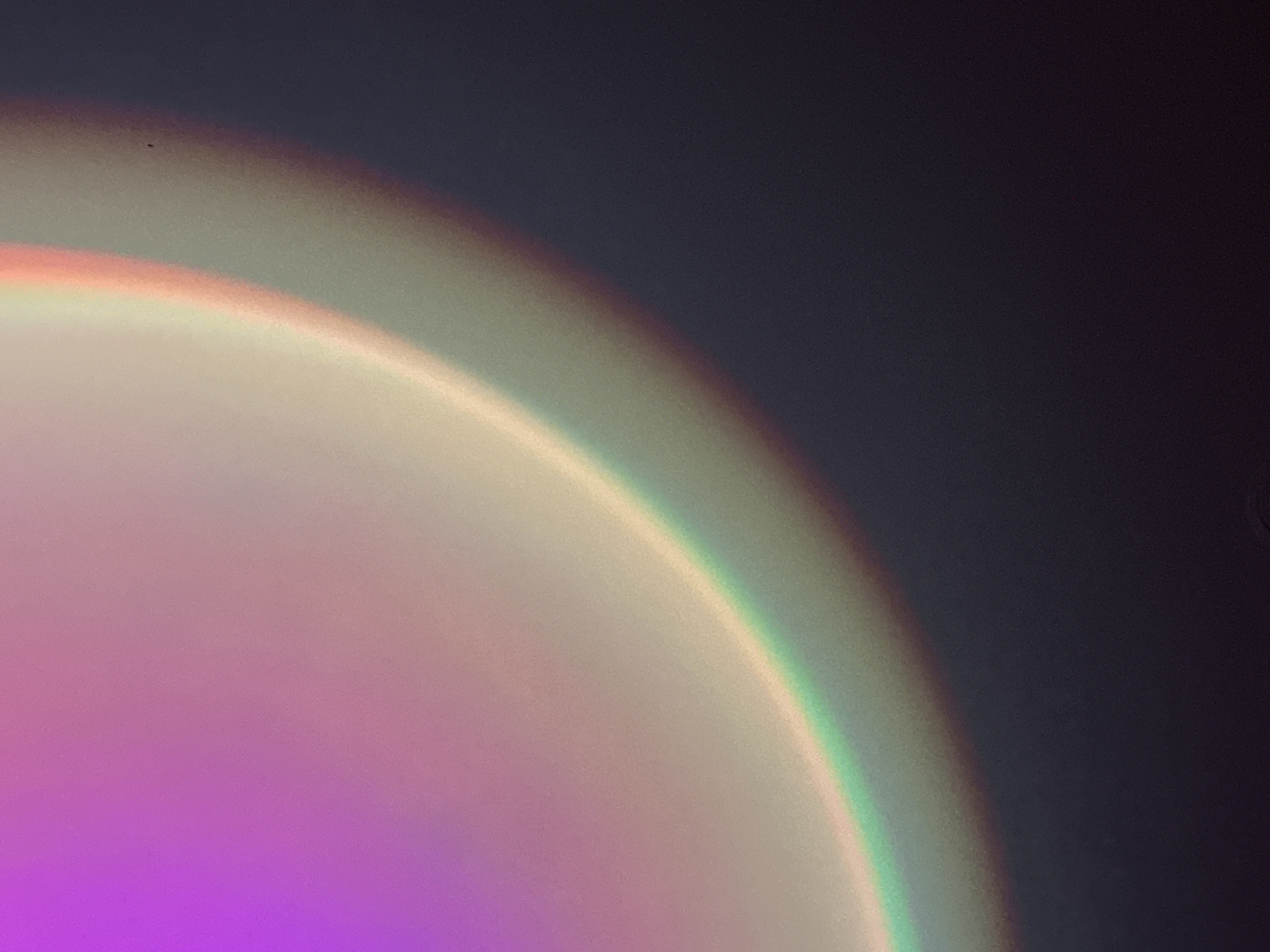 A close-up shot captures a diffused light projection displaying a spectrum of colors. The light emanates from the lower-left corner of the image and curves upwards in a semi-circular arc. The colors blend together, shifting from shades of purple at the bottom to lighter hues of beige and white towards the upper portion of the curve. A distinct band of green and yellow is visible along the edge of the arc, contrasting with the darker gray-blue backdrop that fills the upper-right section of the frame. The photograph has a grainy texture, adding to the soft, hazy quality of the light. The image does not include any indication that it was taken in Chaikovsky, Russia.