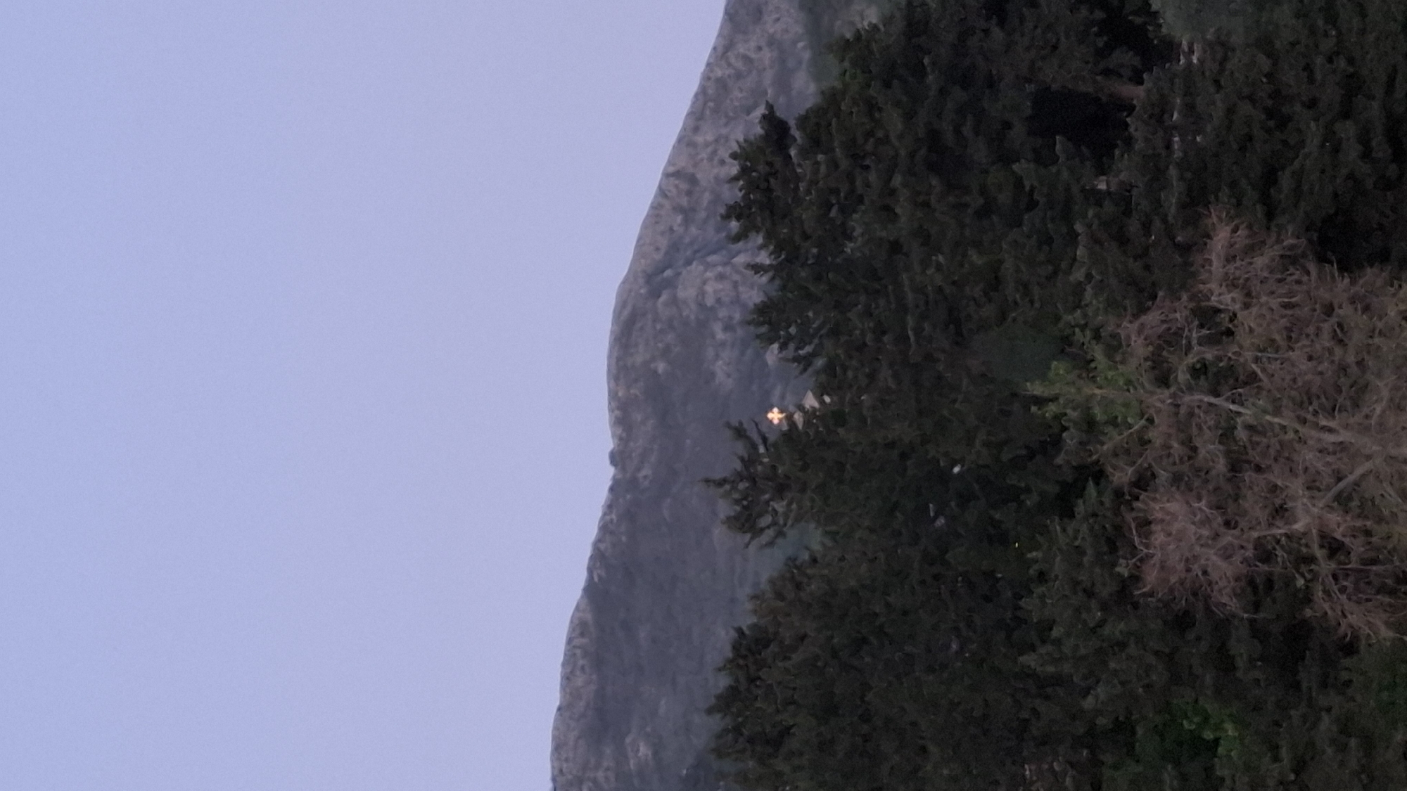 The image shows a mountain in Boreti, Montenegro, next to a treeline. A glowing cross is faintly visible on the side of the mountain. The sky is a pale blue.