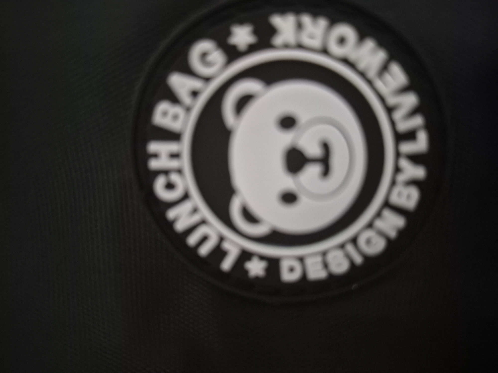 A blurry close-up shot captures a circular emblem featuring a stylized white bear face against a black background. The bear face is simplistic, with rounded shapes defining its eyes, nose, and mouth. The entire image is significantly out of focus, making sharp details difficult to discern.
The circular emblem has text inscribed along its outer edge, set in white against the black rim. Reading clockwise from the bottom left, the text appears to state: "LUNCH BAG * DESIGN * BY LIVE WORK *". Asterisk-like symbols separate the phrases. The words "LUNCH BAG" and "DESIGN" are oriented to be read normally along the bottom arc, while "BY LIVE WORK" is oriented inversely along the top arc.
The emblem is affixed to a dark, possibly black, textured surface, which appears to be a woven fabric, suggesting it might be a bag or a piece of apparel. No specific activity or event is depicted, as the image focuses solely on this static object. There are no discernible location cues, time of day, or weather information.