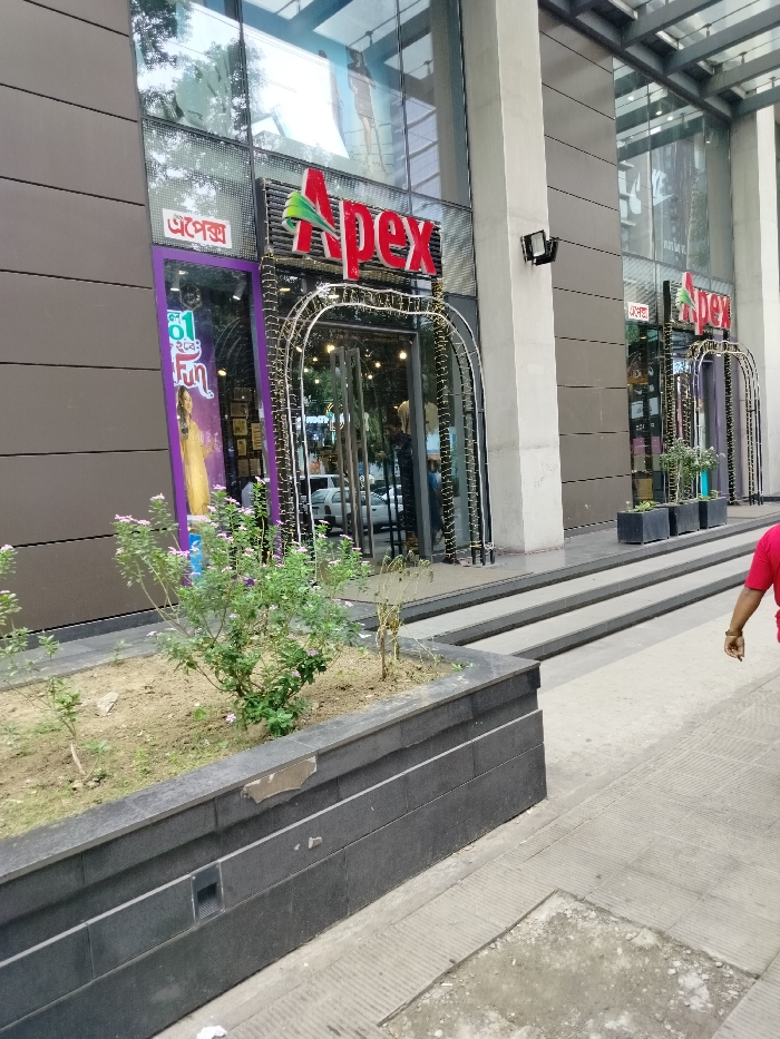 The image shows a storefront for a clothing store called "Apex" located at the coordinates 23.785, 90.417 in Dhaka, Bangladesh. The store is in a modern building with large glass windows and a concrete facade. The storefront is decorated with string lights arranged in arches. There are two large planters in front of the store, one with a bush and the other with a small tree. A single person, wearing a red shirt, is walking past the storefront. The sidewalk is paved with grey tiles. The store has a large sign with "Apex" in red letters and the words "অ্যাপেক্স" (Bengali for Apex) in smaller letters. The storefront window is partially covered with a purple poster, depicting a woman in a sari with the words "Cool Fun" in a bubble. There is a second "Apex" storefront in the background, also with a string light arch. The building appears to be part of a larger shopping complex.