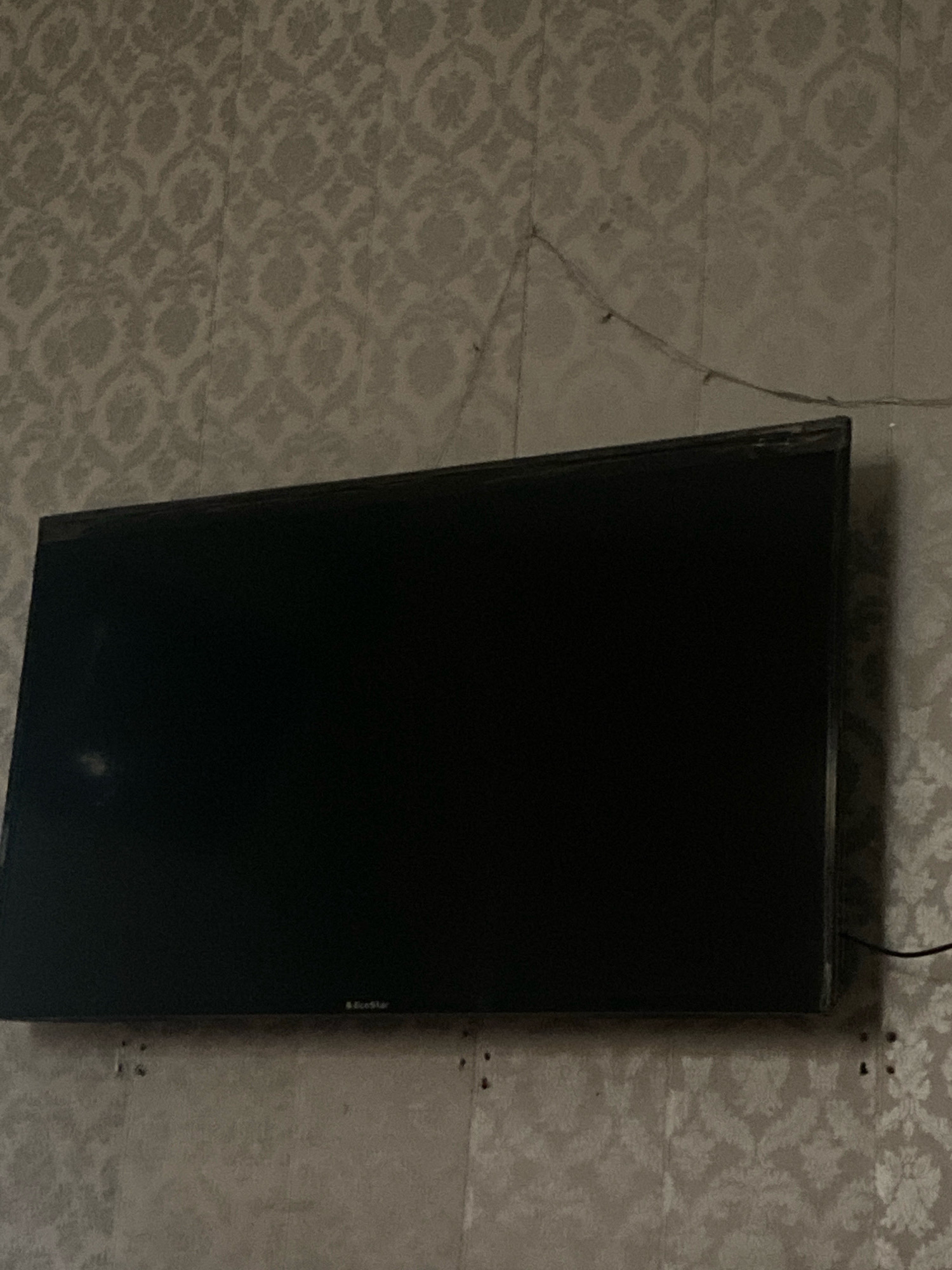 The image shows a flat-screen television mounted on a wall. The wall is covered with patterned wallpaper. The television is black and its screen is dark. There appears to be some dust or smudges on the screen, as well as a faint white mark on the left side. A thin cable is visible running from the top right corner of the television towards the wall. There are also some small holes visible in the wall below the television, which may indicate mounting hardware. The lighting in the image is dim, casting shadows and making it difficult to discern finer details of the wallpaper pattern. This scene is consistent with a domestic setting within a home in Gujranwala City Tehsil, Pakistan, where such televisions are commonly found mounted on walls.