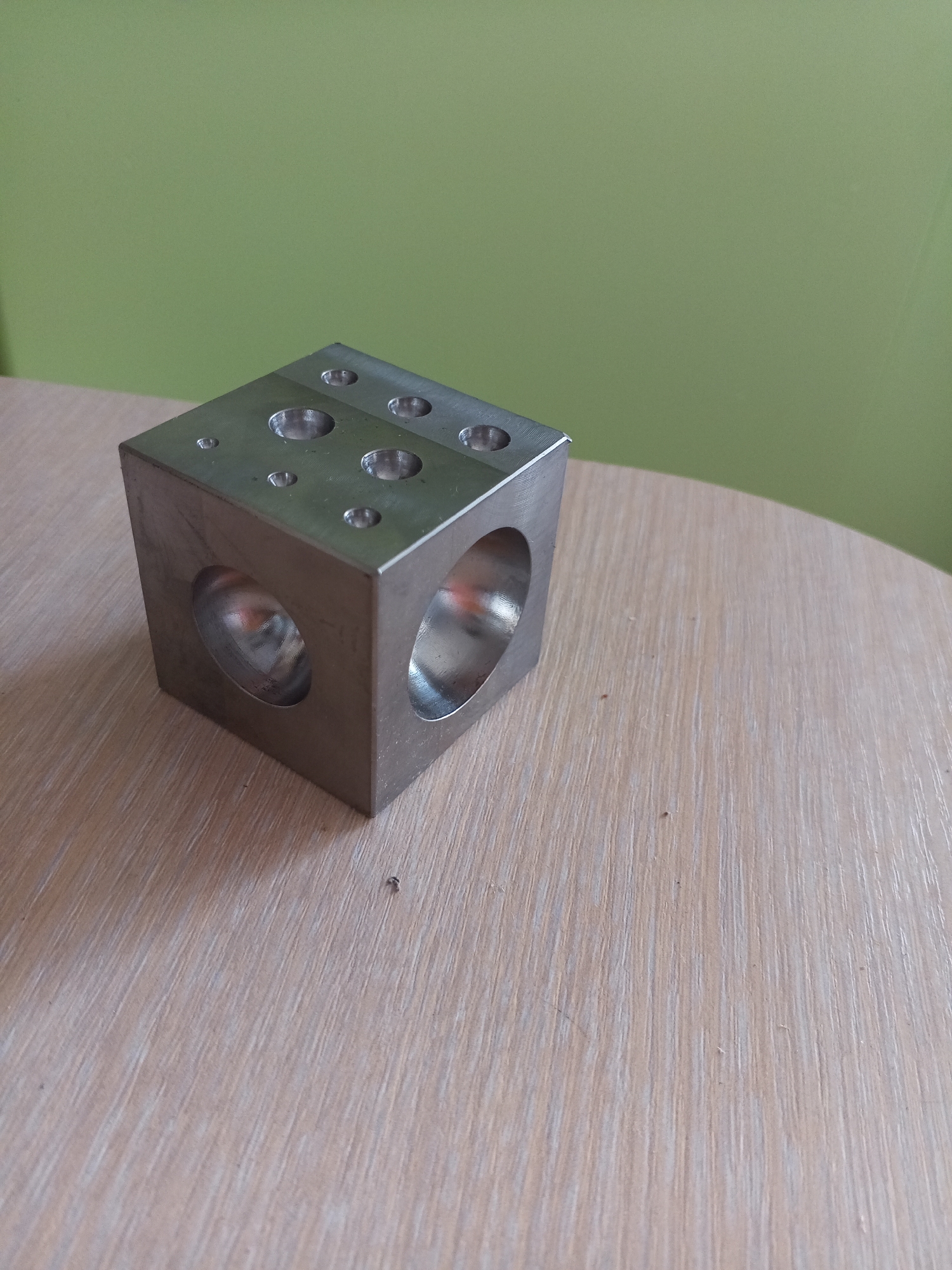 A close-up, slightly elevated shot shows a metal cube with circular holes on its top and sides, resting on a light-colored, wood-grained surface. The cube is positioned to the left of the frame, with its top surface facing the camera and slightly angled. Three circular indentations are visible on the top surface, varying in size. Two large, circular openings are visible on the front and right sides of the cube, revealing reflections within their hollow interiors. The background consists of a solid, muted green wall. The lighting appears to be natural or ambient, casting soft shadows. The image is crisp and in focus, emphasizing the texture of both the metal cube and the wooden surface. The scene is devoid of any human presence or activity. The location of Vilyuysk, Russia, is not ascertainable from the visual information provided in the image.
