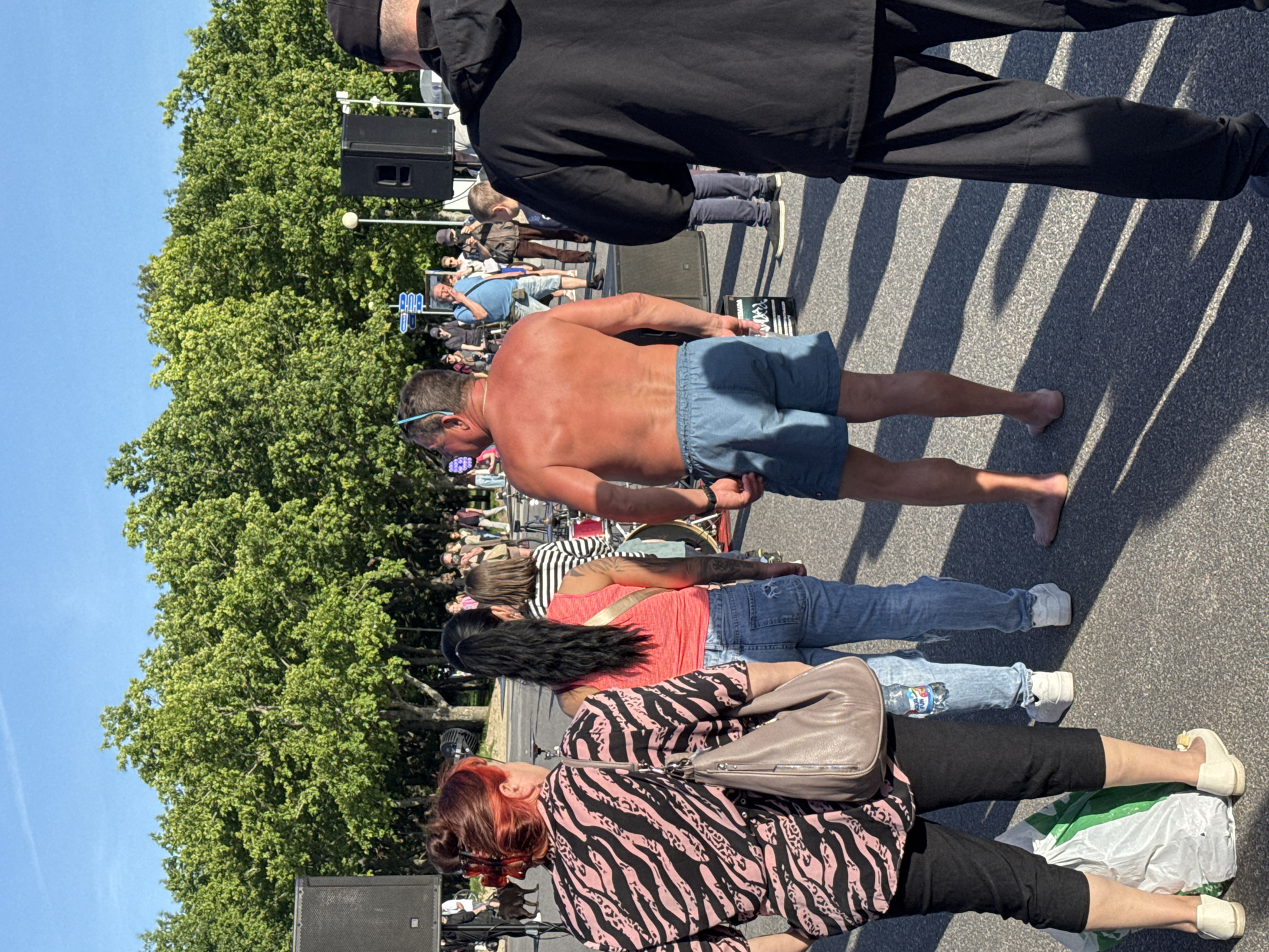 The media file depicts a sunny outdoor scene, likely in Tallinn, Estonia, with people gathered in front of a stage or performance area. The time of day appears to be late morning or afternoon due to the strong shadows cast by the sun.
In the foreground, a man with a tanned, shirtless torso and blue shorts is prominently featured. Behind him, several people are standing. A woman in a red tank top and jeans, another with dark hair and a striped top, and a third with distinctive reddish hair and a black and pink zebra-print top are visible. One woman is carrying a light brown purse. A water bottle is visible in the back pocket of a woman's jeans.
Further back, a crowd of people can be seen, some standing and some appearing to be seated or leaning. Large speakers are positioned in the background, suggesting a concert, festival, or public event. Trees with lush green foliage line the area, indicating a pleasant, possibly warm, day. The ground is asphalt, marked by distinct shadows, reinforcing the sunny weather. The overall atmosphere seems relaxed and social, with people enjoying an outdoor gathering. No specific text is clearly legible in the image.