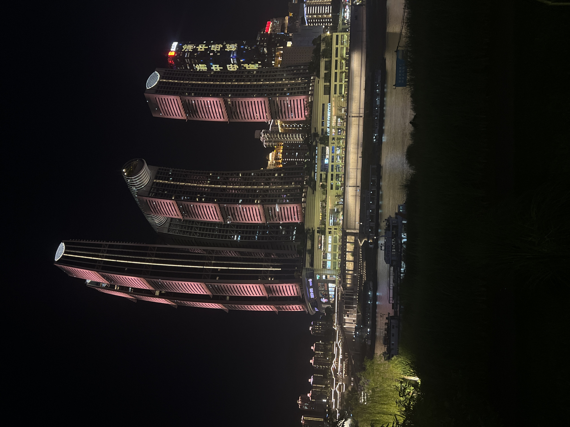 The image captures a nighttime scene of modern architecture and a waterfront. Three prominent, illuminated skyscrapers with curved facades and pink striped detailing dominate the left side of the frame. These structures are lit from within and by external lights, highlighting their sleek, contemporary design. Visible Chinese text, appearing as bright yellow characters on one of the buildings, suggests the location is in China, specifically within the Jiangbei Cheng Subdistrict.
To the right of the skyscrapers, a series of lower buildings are visible, also illuminated. A pathway or road runs alongside these structures, bordered by what appears to be water reflecting the lights. Further to the right, a body of water is present, with several boats or barges moored along the bank. The dark sky indicates it is nighttime. The overall atmosphere is one of urban development and nighttime activity. The focus is on the architecture and the illuminated cityscape, with no discernible people or specific events taking place. The presence of water and the style of the buildings suggest a developed urban waterfront area.