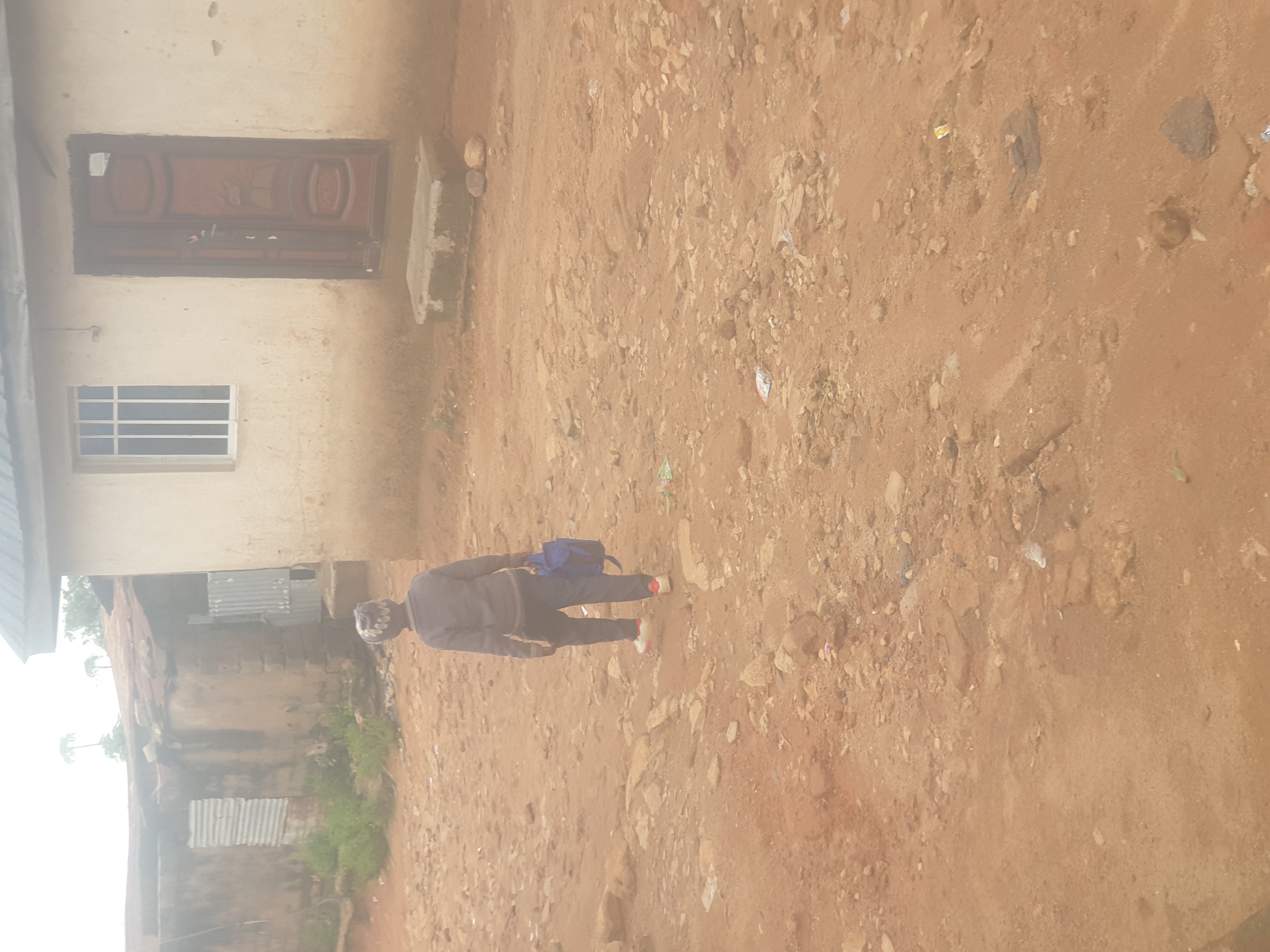 The image shows a person wearing a gray sweatshirt, dark pants, a patterned cap, and white sneakers with red accents, walking away from the camera on a dirt path. The person is carrying a blue backpack in their right hand. The ground is covered in reddish-brown dirt and scattered rocks. In the background, there is a light-colored building with a dark door and a window with white bars. To the left of the building, there is another structure with a corrugated metal roof and walls, and some green foliage. The overall environment appears to be rural or a less developed urban area. There is no identifiable information to indicate the location is in Washington, United States of America.
