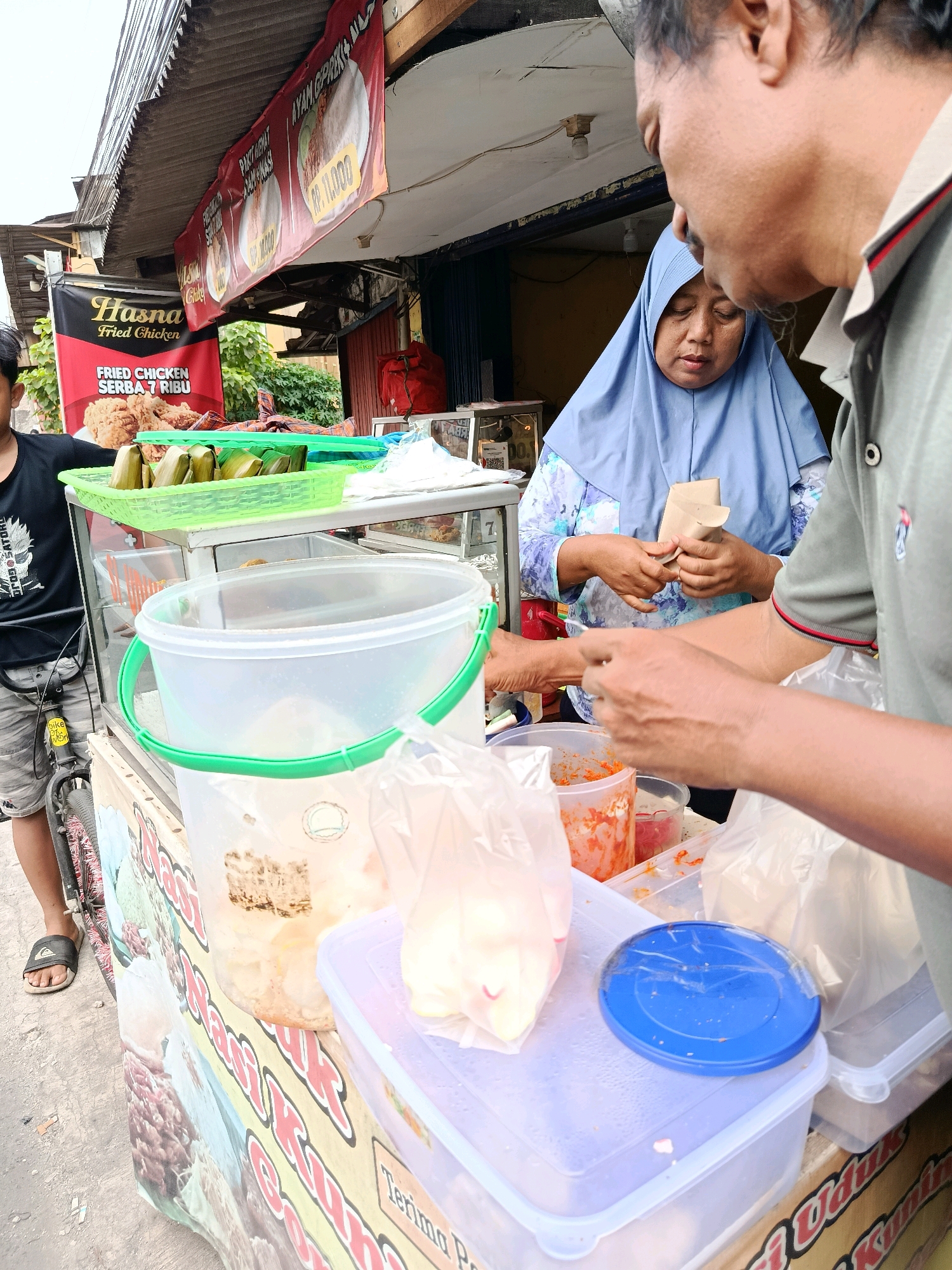 The image depicts a street food stall in Bekasi, Indonesia. Two individuals, a woman wearing a blue hijab and a man, are interacting at the stall. The woman is holding a small brown paper package and appears to be receiving it from the man, who is in the foreground with his back to the camera. The stall is laden with food items, including what appear to be pre-packaged meals wrapped in banana leaves in a green basket. Several plastic containers hold various food ingredients, possibly sambal or other condiments, one of which is red and finely chopped. A large white plastic bucket with a green handle is also visible. In the background, banners advertise "Hasna Fried Chicken" with prices and other food items. A portion of a bicycle is visible to the left, with a person's legs and feet wearing sandals. The overall environment suggests a busy outdoor market or roadside vendor setting.