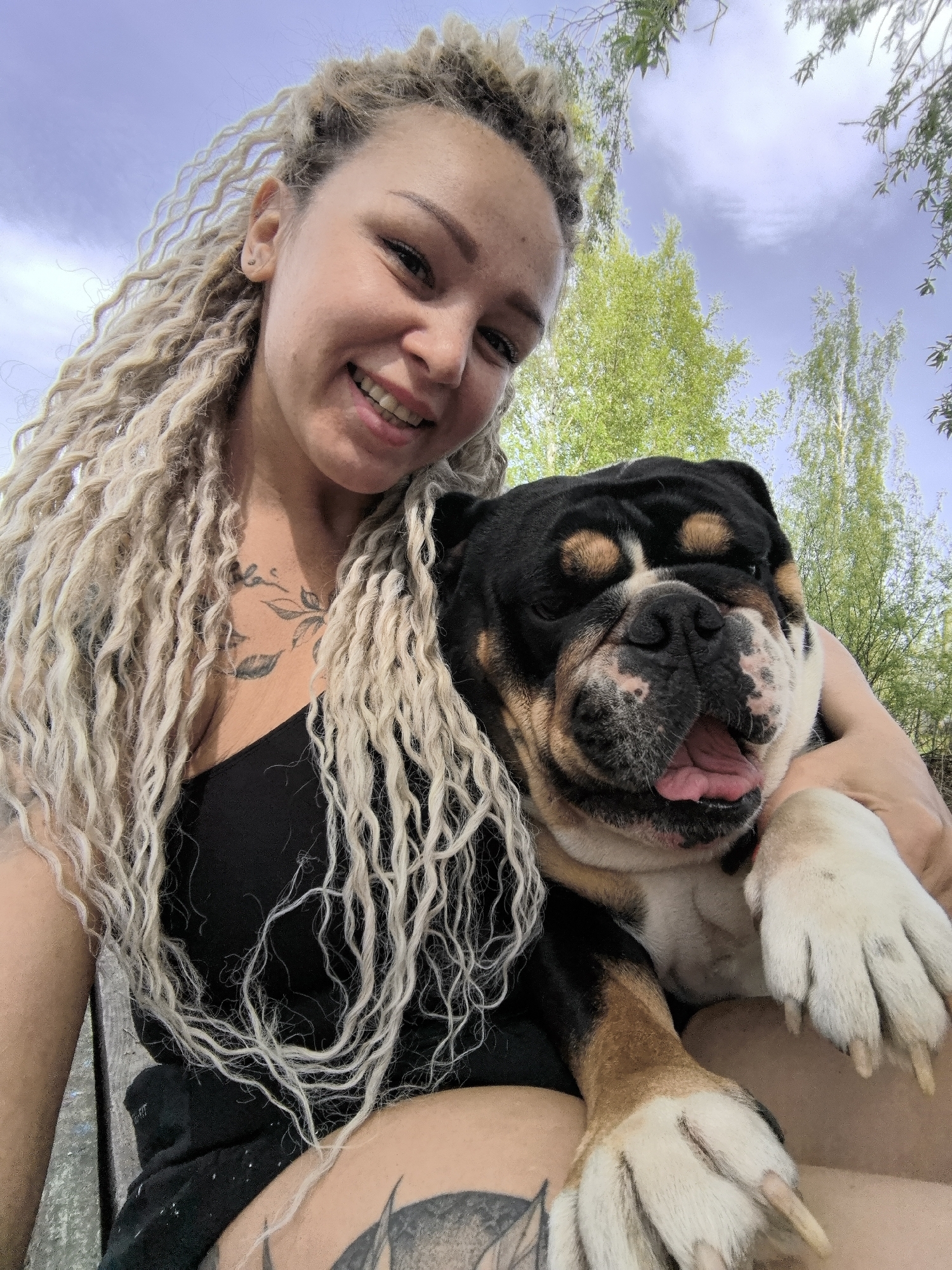 A woman with blonde dreadlocks is holding a black and brown English Bulldog. The woman is smiling and wearing a black tank top. She has a tattoo on her chest and a tattoo on her thigh. The dog has a white and brown face, and its tongue is sticking out. The background includes green trees and a blue sky with clouds. The image was likely taken outdoors. The exact location within the city of Noviy, Russia, cannot be determined from the provided information.
