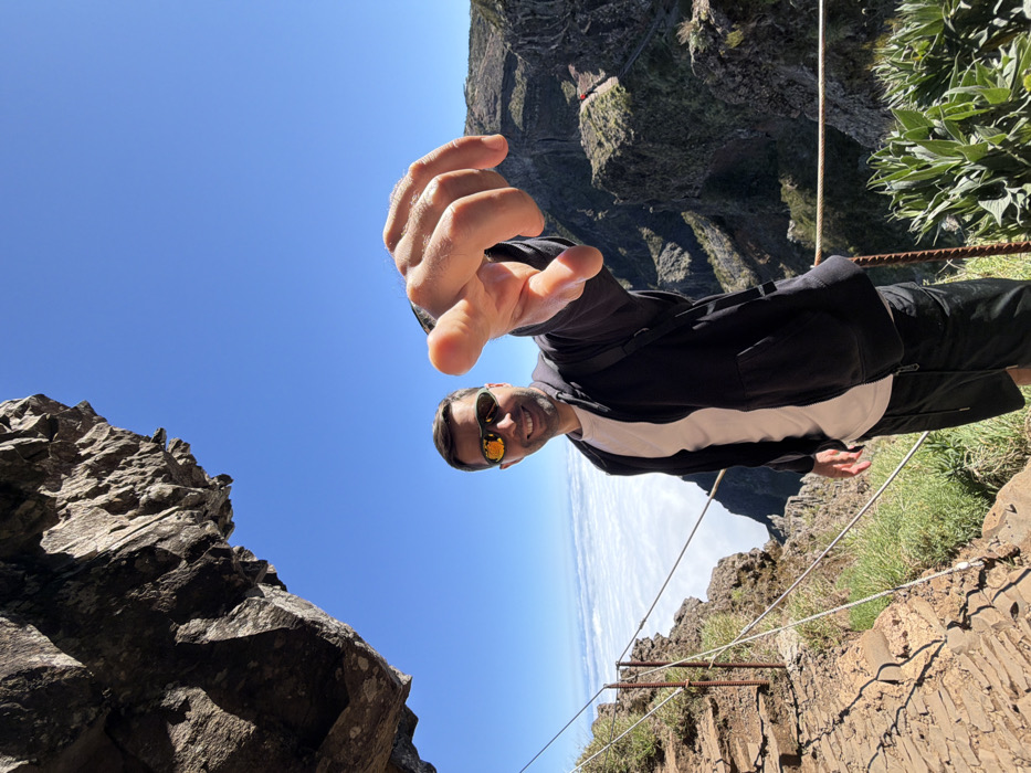The image captures a male subject on a high-altitude mountain hiking trail, consistent with the mountainous interior of Madeira, Portugal, within the broader region of Câmara de Lobos. The subject, an adult male with short dark hair and a beard, is positioned slightly off-center to the left, smiling directly at the viewer. He wears dark reflective sunglasses, a light-colored t-shirt, and an unzipped black hooded jacket over dark shorts. His right arm is extended towards the camera, with his hand open and index finger pointing forward in a direct gesture towards the viewer.
To the subject's left is a narrow, stony path lined with a multi-strand wire safety railing secured by metal posts. An overhanging dark rock formation is visible in the upper left. Below the path and extending into the background is a dense layer of white clouds, indicating a cloud inversion. Jagged, dark green and grey mountain ridges and peaks emerge from or rise above these clouds. A distant winding trail with a small, red-clad figure (another hiker) is discernible on a ridge in the far right background. The sky above is clear and deep blue. Sparse green vegetation grows on the immediate rocky slopes. The scene is illuminated by bright daylight.