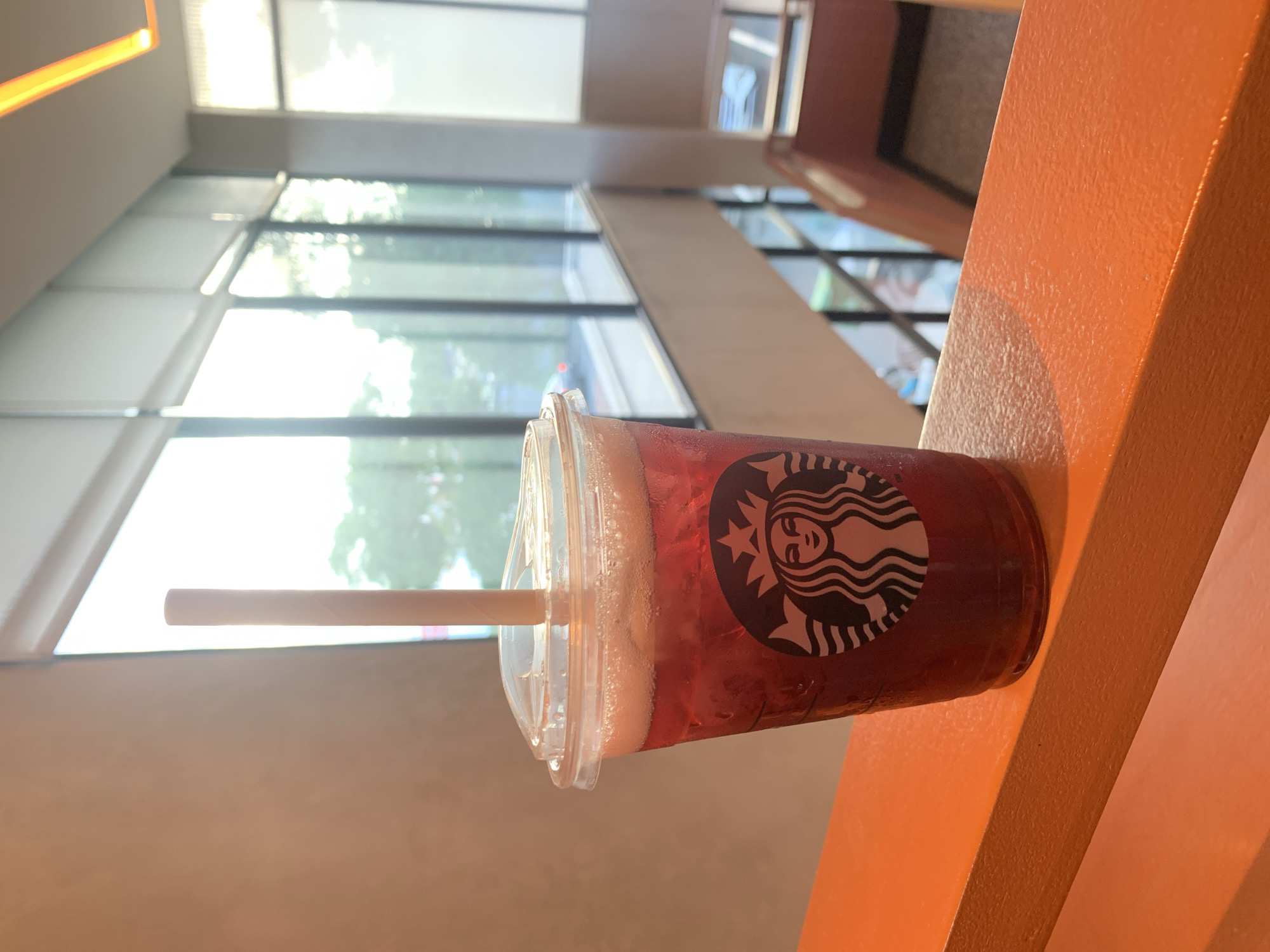 A cold Starbucks beverage in a clear plastic cup with a straw sits on an orange railing. The cup has the Starbucks logo visible. The background shows a brightly lit indoor space with large windows overlooking an outdoor area with trees and what appears to be a city street. The railing is smooth and painted orange. The lighting suggests it is daytime. There are no people in the frame. This scene could be a cafe or a seating area within a building, possibly in a commercial district in Angeles City, Philippines, given the architectural style and general urban environment suggested by the background.