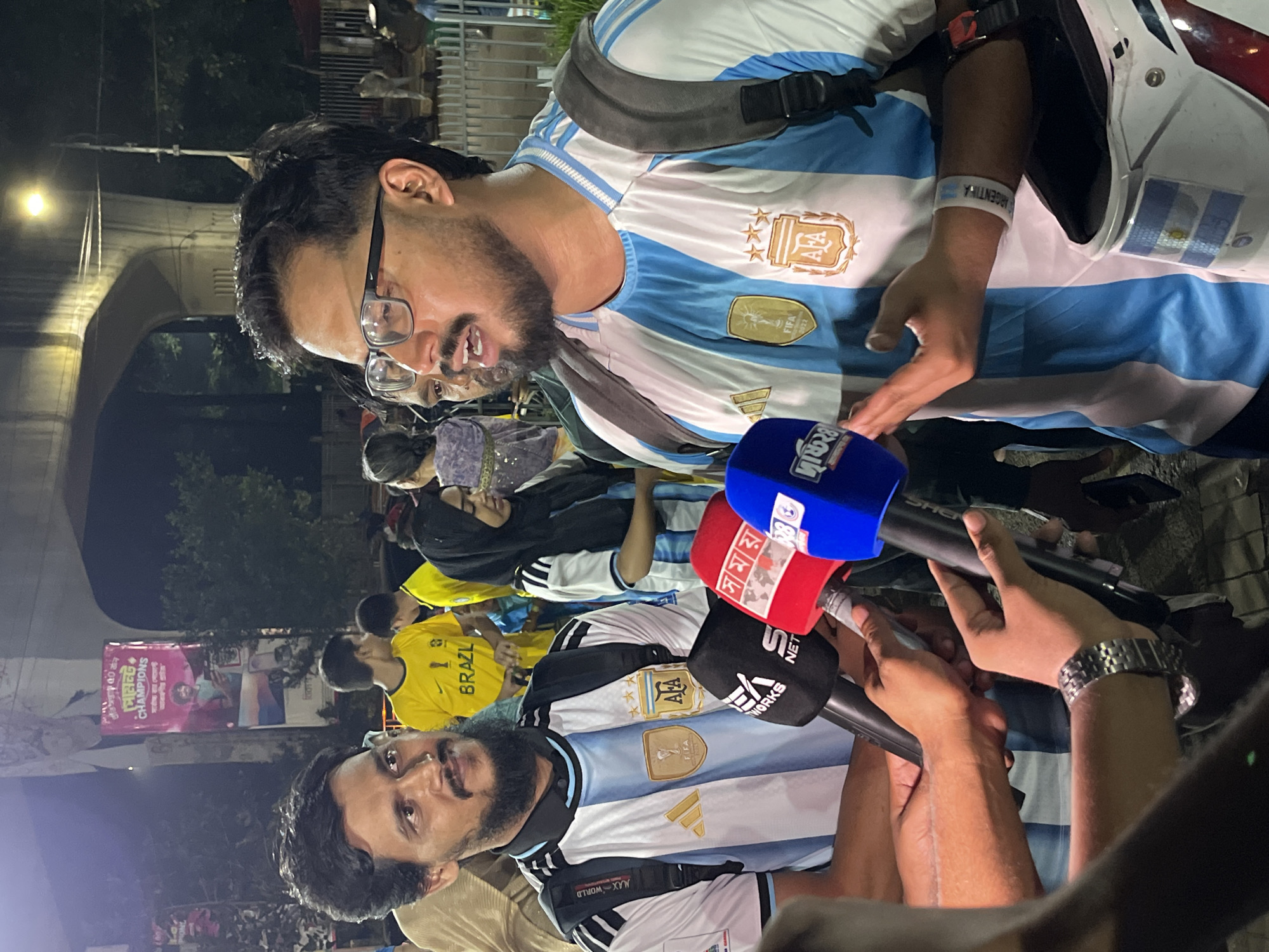 In Dhaka, Bangladesh, on a street at night, a man wearing an Argentina jersey and a backpack is being interviewed by several reporters holding microphones. The man has a beard and glasses and is speaking animatedly. Behind him, other people in similar jerseys are visible, along with a large banner depicting a man and text in Bengali. The scene appears to be a public gathering, possibly related to a sporting event.