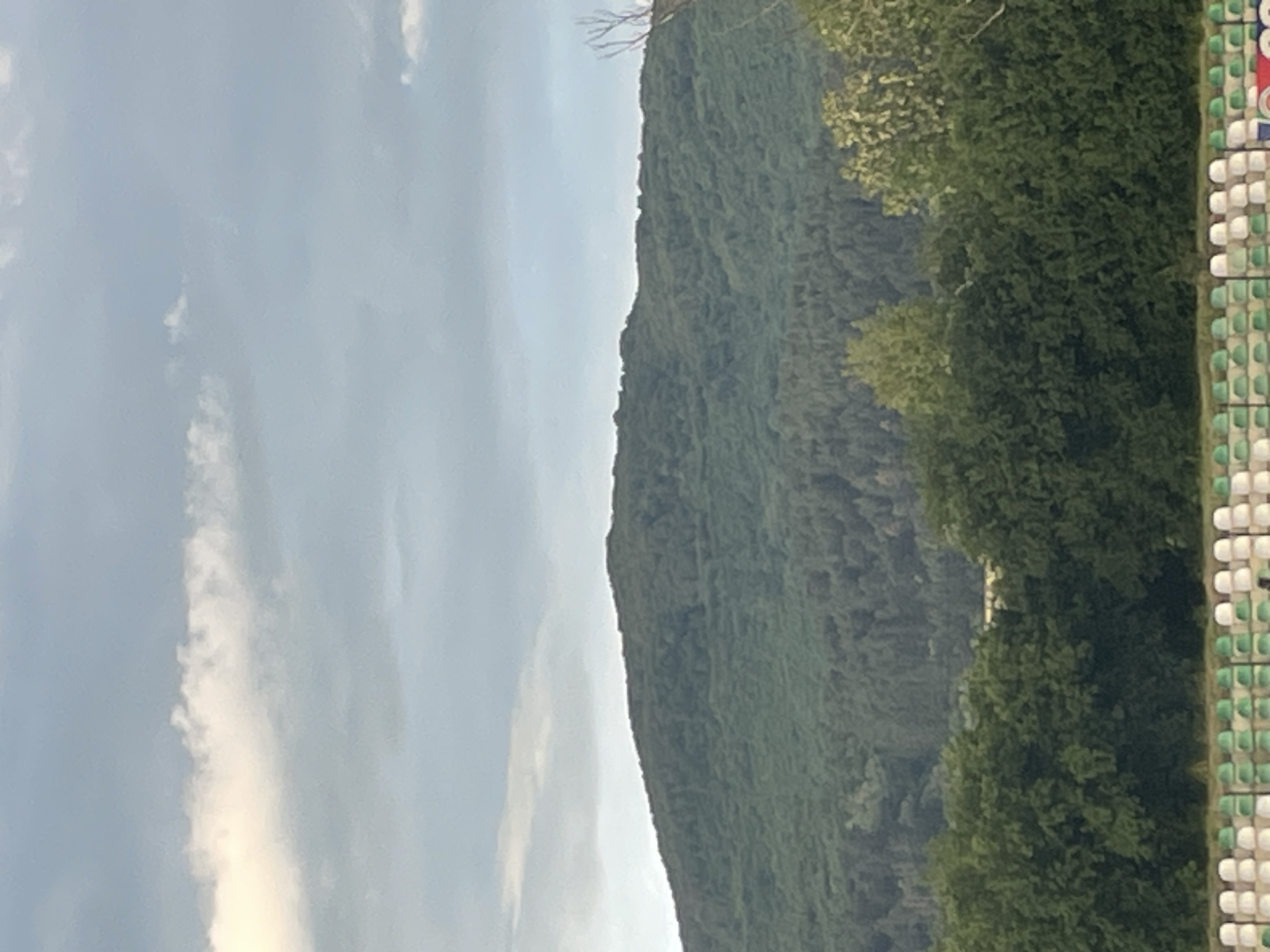 The image displays a daytime landscape featuring a large, densely forested mountain or hill against a partially cloudy, light blue sky. The mountain, covered in various shades of green foliage, occupies the central and left parts of the frame, with its irregular silhouette forming a clear horizon line. On the far right of the image, a section of outdoor stadium seating is visible. The seats are arranged in orderly rows and columns, primarily in alternating white and green colors, with a few red accents discernible. The foreground consists of trees, appearing brighter green than the distant mountain foliage. The weather appears fair with scattered white clouds. No people or specific activities are visible, and there is no discernible text. The scene depicts a natural setting adjacent to an artificial structure, suggesting a sports facility or arena nestled within a mountainous environment, consistent with the location context of Ihtiman, Bulgaria.