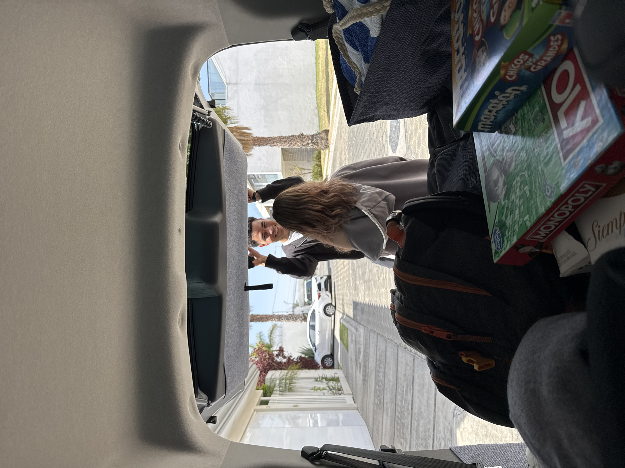The view is from inside the open hatchback of a vehicle. In the cargo area are items including a backpack, towels, and board games like Monopoly, OK, and a Marathon game. A woman and a man are standing outside the vehicle, near the back. They appear to be interacting with something inside the trunk. The setting is a residential street in San Bernardino Tlaxcalancingo, Mexico, as evidenced by the architecture and palm trees. There is a white vehicle parked on the street outside the house.