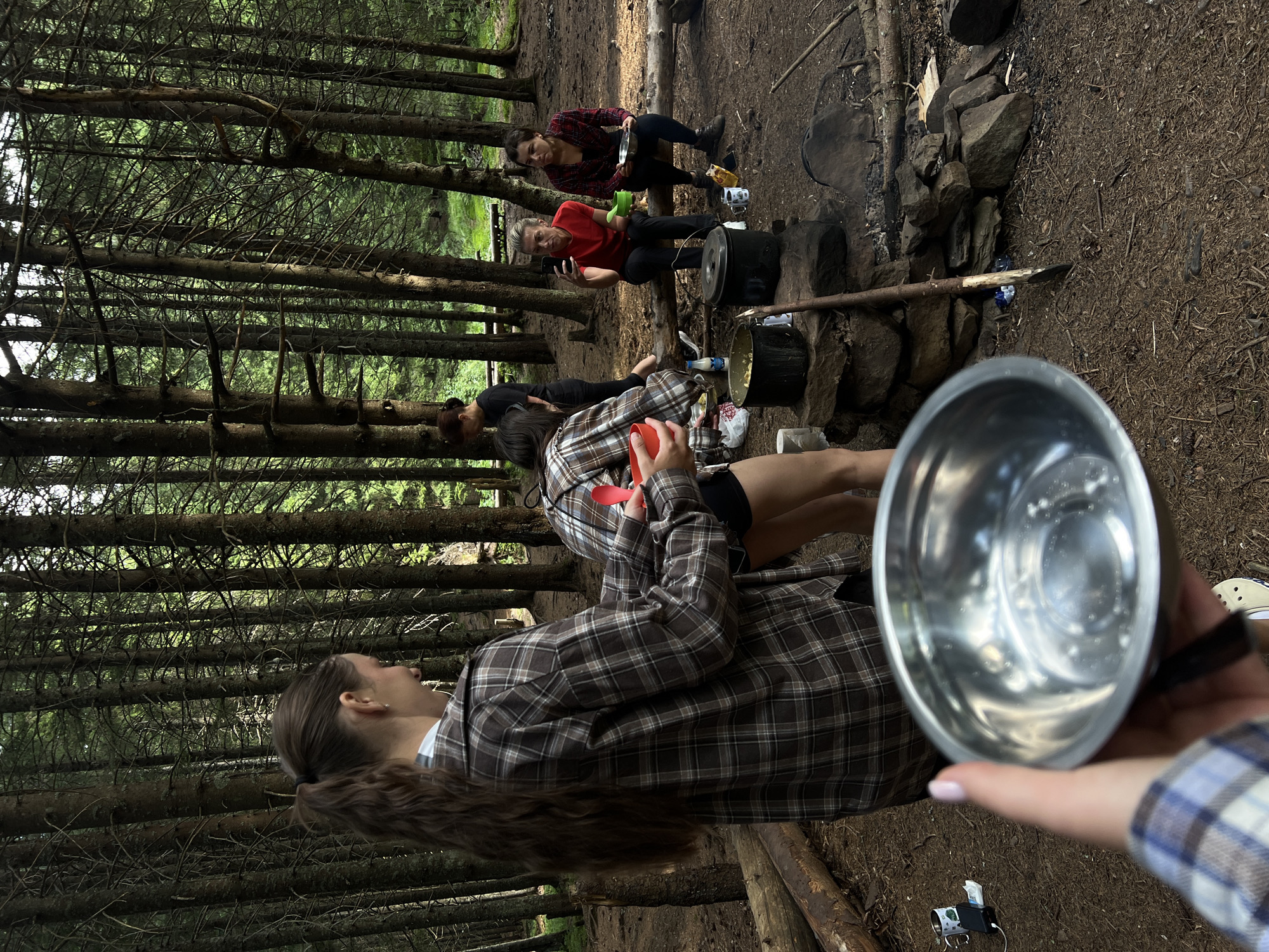 In a dense coniferous forest in the city of Vorokhta, Ukraine, a group of individuals is gathered around a makeshift campfire. The forest floor is covered with pine needles and scattered branches. In the foreground, a person's hand holds a silver bowl, partially obscuring the view of the scene.
Further into the clearing, two women are tending to cooking pots situated on a stone structure that serves as a hearth. One woman, wearing a plaid shirt, is actively stirring the contents of a pot, while another, also in a plaid shirt, holds a red bowl.
In the background, two other individuals are seated on a fallen log. One woman, wearing a red top, is looking at her phone, and the other, in a plaid shirt, is holding a bowl. Another person, wearing a dark top, is standing behind them. The overall atmosphere suggests a casual outdoor gathering or camping trip. The trees, characteristic of the Carpathian Mountains region, create a natural, secluded setting.