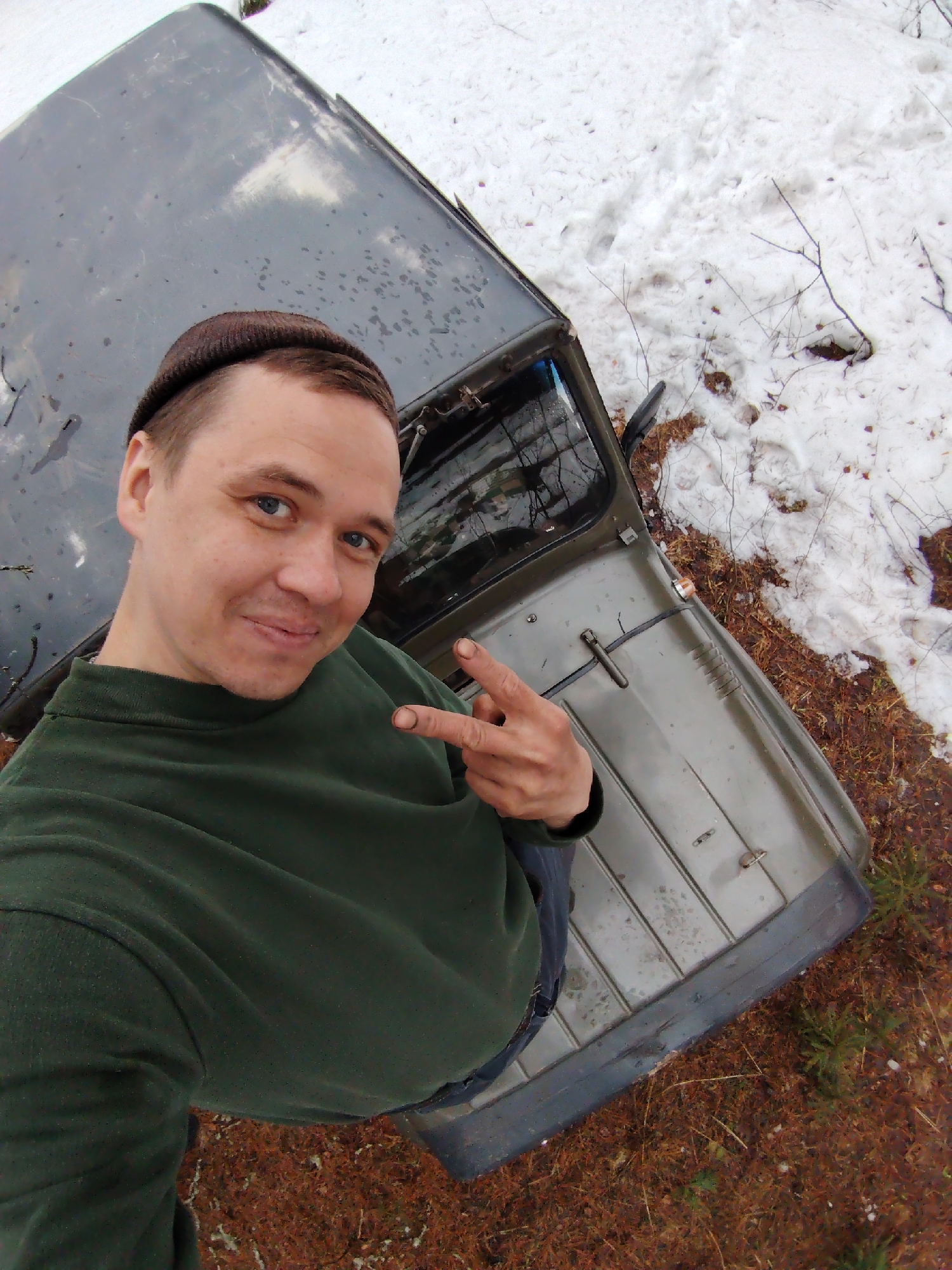 The image is a high-angle selfie of a man taken outdoors, with an olive green off-road vehicle and a snowy, wooded landscape in the background. The man, with short light brown hair and blue eyes, wears a dark brown beanie and a dark green long-sleeved shirt. He is smiling faintly at the camera, holding up his right hand in a "peace" or "victory" sign. His fingers appear smudged with dirt or grease.
The vehicle, possibly a rugged SUV or jeep, shows signs of use with smudges on its dark roof and hood. Part of the driver's side door and a side mirror are visible. The ground is covered in patches of white snow and reddish-brown earth mixed with pine needles or dry foliage, typical of a forest or rural area in Markovskii, Russia. Sparse, bare branches are visible on the ground. The presence of snow suggests a cold climate, likely late winter or early spring. The lighting is natural and diffused, indicating daytime, possibly under an overcast sky. The overall scene conveys a casual, rugged, and adventurous outdoor setting. No visible text is present.