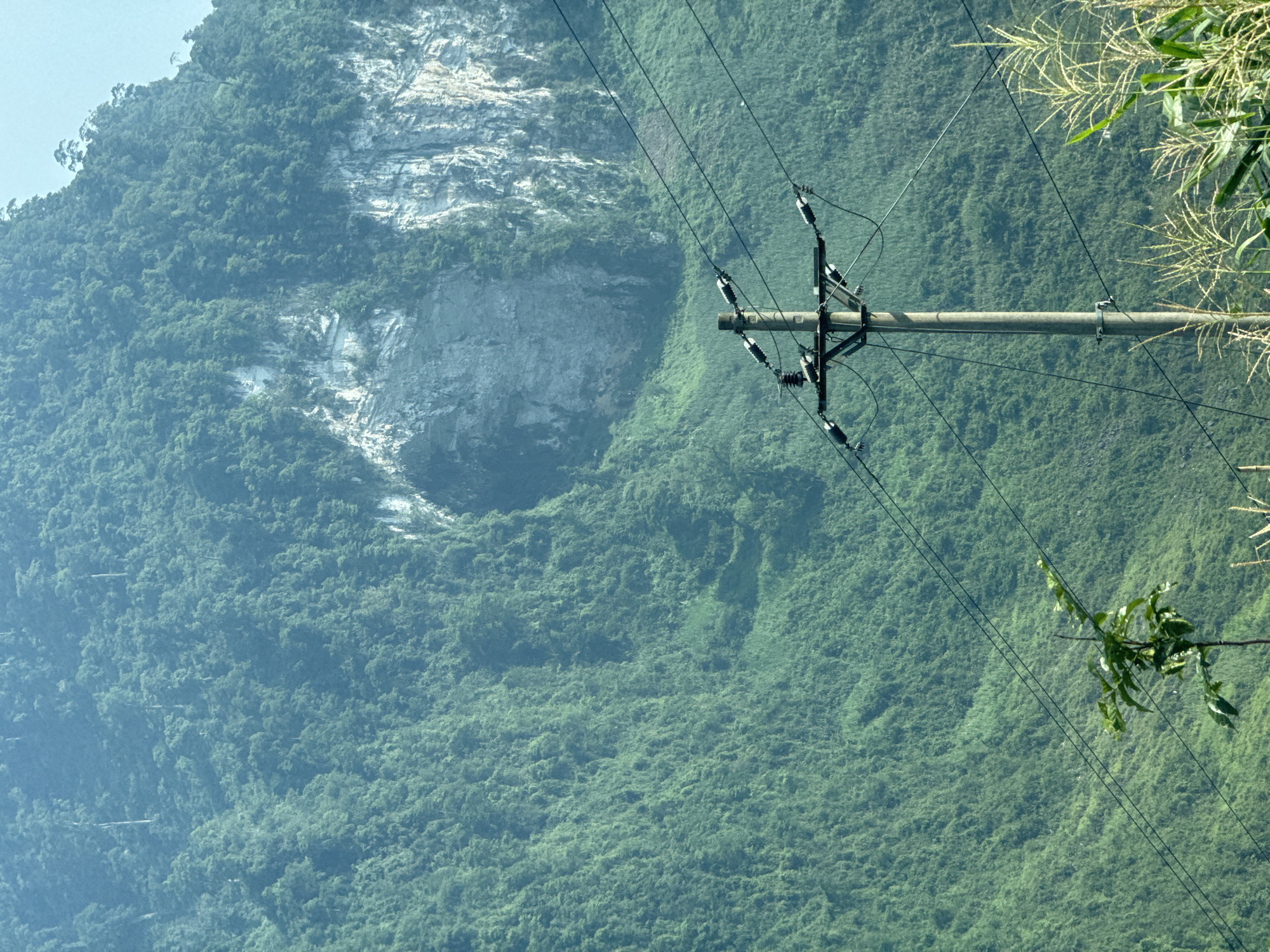 The image shows a lush, green, mountainous landscape with a prominent grey rock face. A utility pole with power lines crosses the view, with foliage from nearby plants obscuring the upper right corner and a branch extending from the right toward the lower-center area. The photo seems to be taken in or near Đường Thượng Commune, Vietnam given its mountainous terrain and dense vegetation.