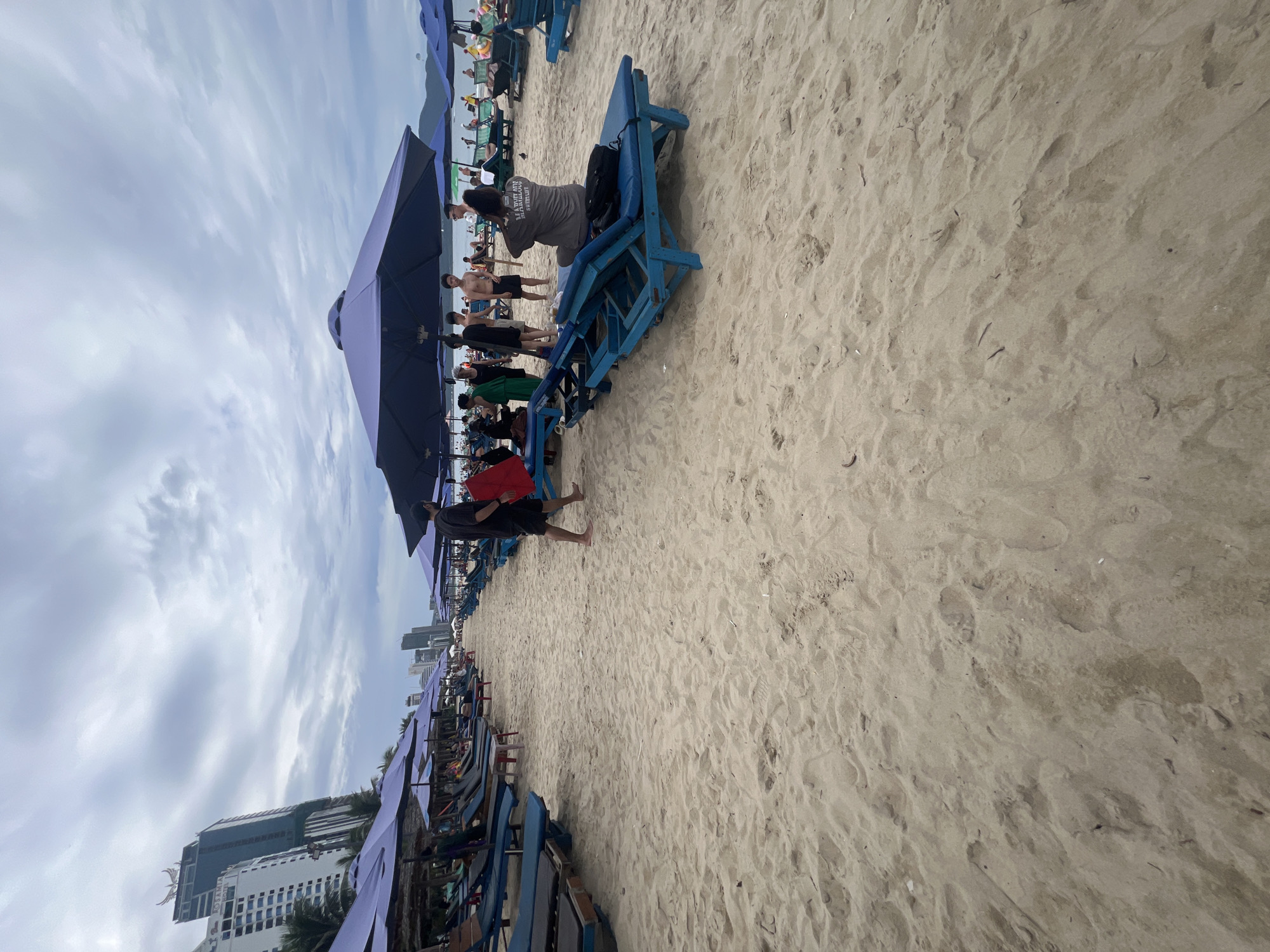 The image depicts a crowded beach scene in Đà Nẵng, Vietnam, under an overcast sky. The foreground is dominated by light-colored sand, textured with footprints and debris. Numerous beach umbrellas, mostly purple, are set up in rows, providing shade. Blue beach chairs are scattered amongst them, some occupied, others empty.
In the middle ground, several people are visible. A group of young men, some shirtless, are gathered near an umbrella. One man in a grey t-shirt with text, possibly a logo or slogan, is sitting on a blue beach chair, holding a black backpack. Another person in the foreground is wearing a red shirt and black shorts. The people appear to be engaging in casual beach activities, with some standing and conversing, while others are seated.
In the background, tall buildings and hotels line the coastline, indicating an urban setting adjacent to the beach. The sky is cloudy, suggesting it might be morning or late afternoon, or that the weather is generally grey. The overall atmosphere seems to be one of a busy public beach. No specific event is clearly identifiable, but it appears to be a typical day with people enjoying the beach facilities.