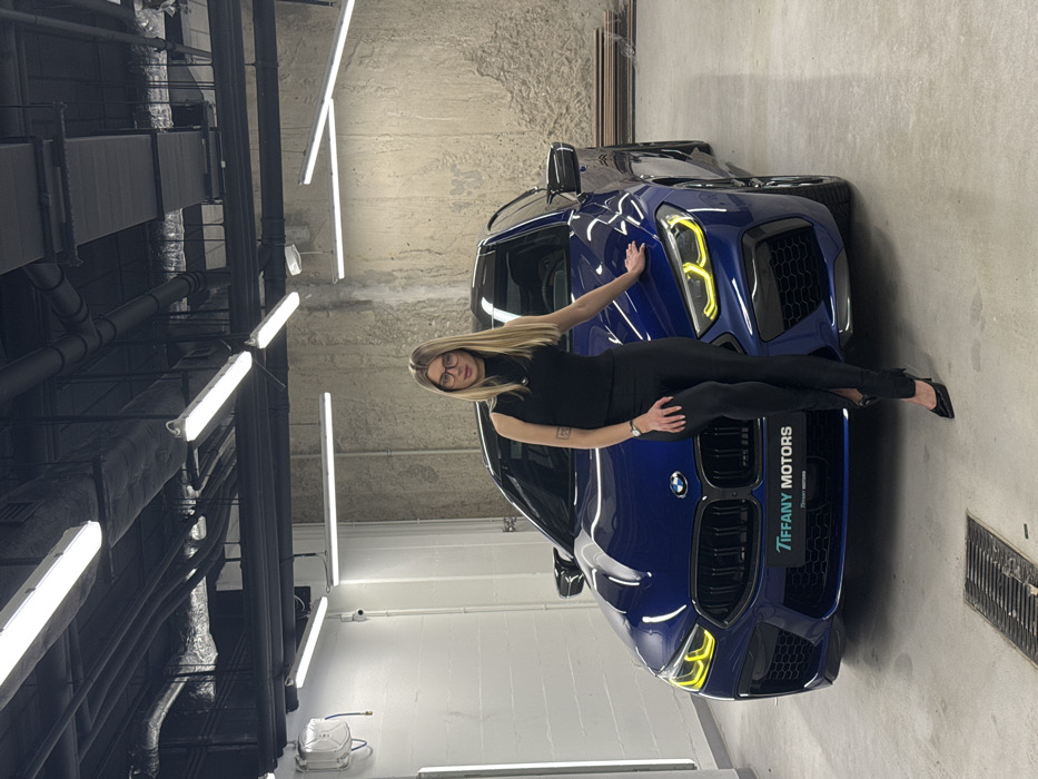 A woman with blonde hair and glasses lies across the hood of a blue BMW with a "Tiffany Motors" license plate. She is wearing a black top and pants with black heels. The setting appears to be a garage or showroom with industrial lighting fixtures on the ceiling. Another dark car is partially visible behind the blue one.