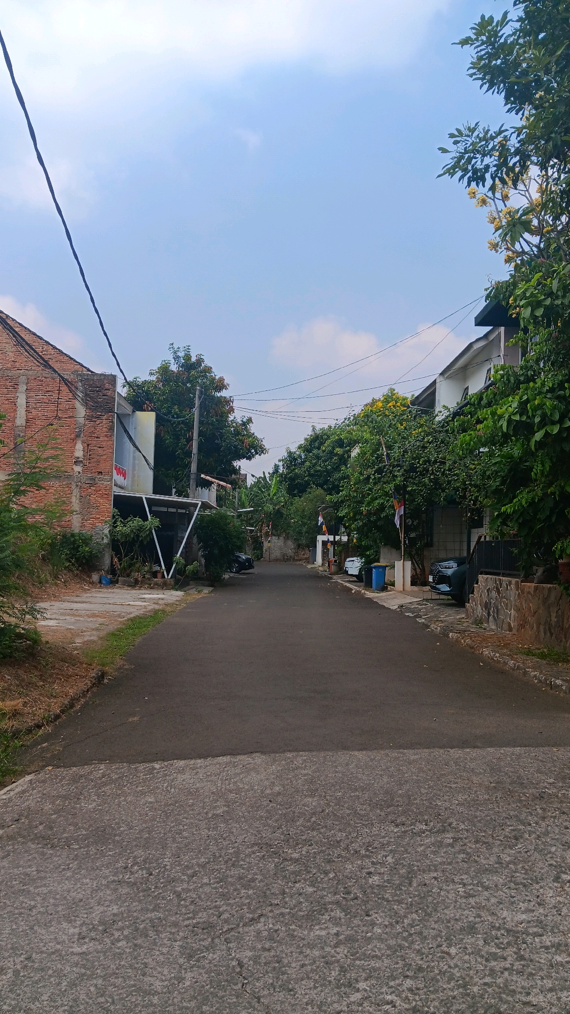The image depicts a quiet residential street in Depok, Indonesia. The street is paved with asphalt and concrete. On the left side, a brick building with visible repairs stands next to a more modern house with a covered carport. Several potted plants are arranged along the driveway. On the right side of the street, there are modern houses with lush green trees and flowering plants. Two cars, a dark SUV and a smaller vehicle, are parked in driveways. A blue trash bin is also visible on the right. Several Indonesian flags are displayed on poles in front of some of the houses. The sky is overcast with a light blue hue. Overhead power lines are visible, stretching across the street. The overall scene suggests a peaceful, ordinary day in a suburban neighborhood.
