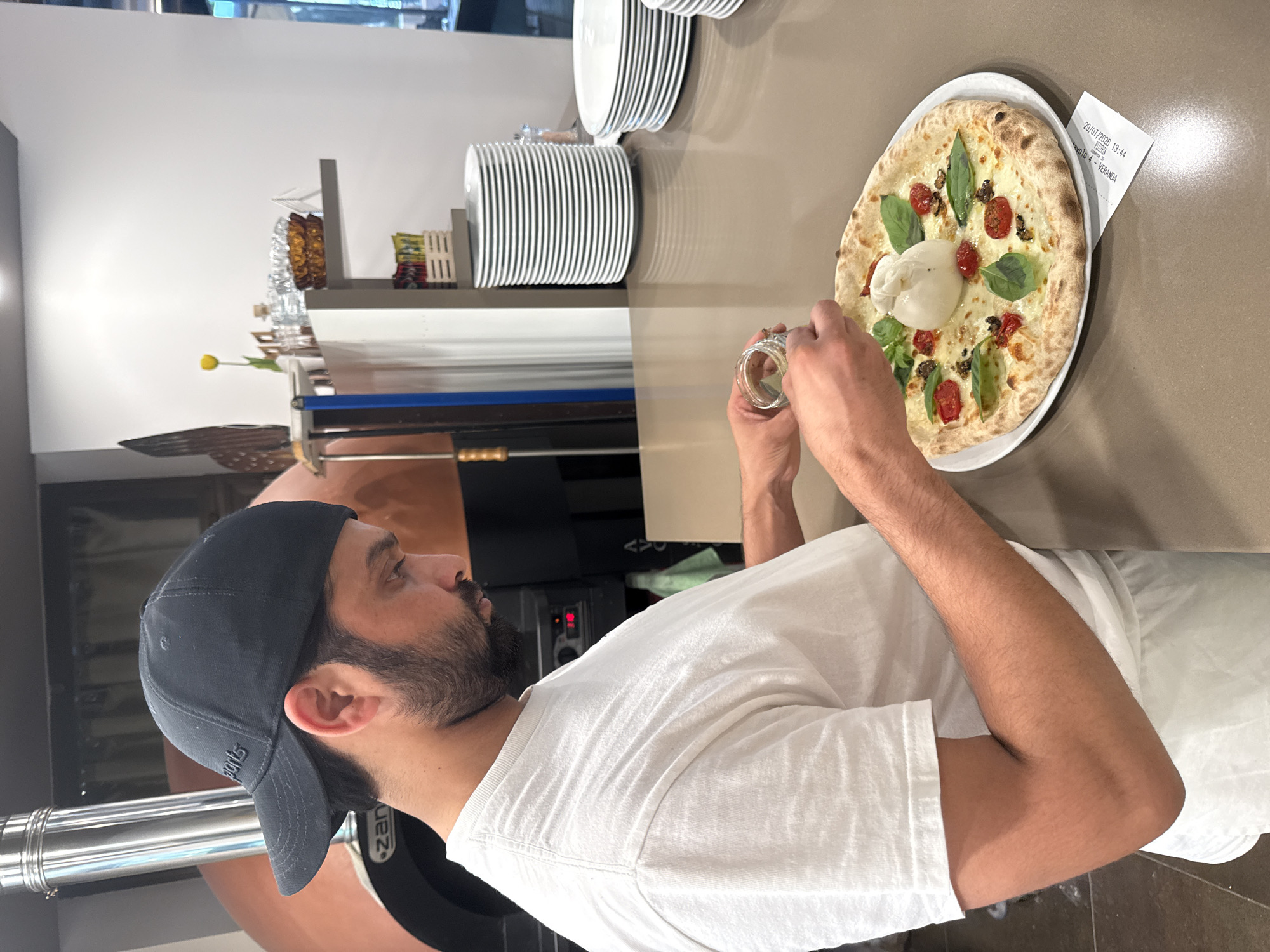 The image shows a man wearing a black baseball cap and a white t-shirt, preparing a pizza. He is holding a small jar and sprinkling something onto the pizza, which is topped with cheese, tomatoes, basil, and a large ball of burrata cheese. The pizza is on a white plate on a counter. In the background, there is a pizza oven, stacks of plates, and shelving with various items. The receipt under the pizza indicates the location is Trento, Italy, with a date and time of 29/07/2025 13:44.